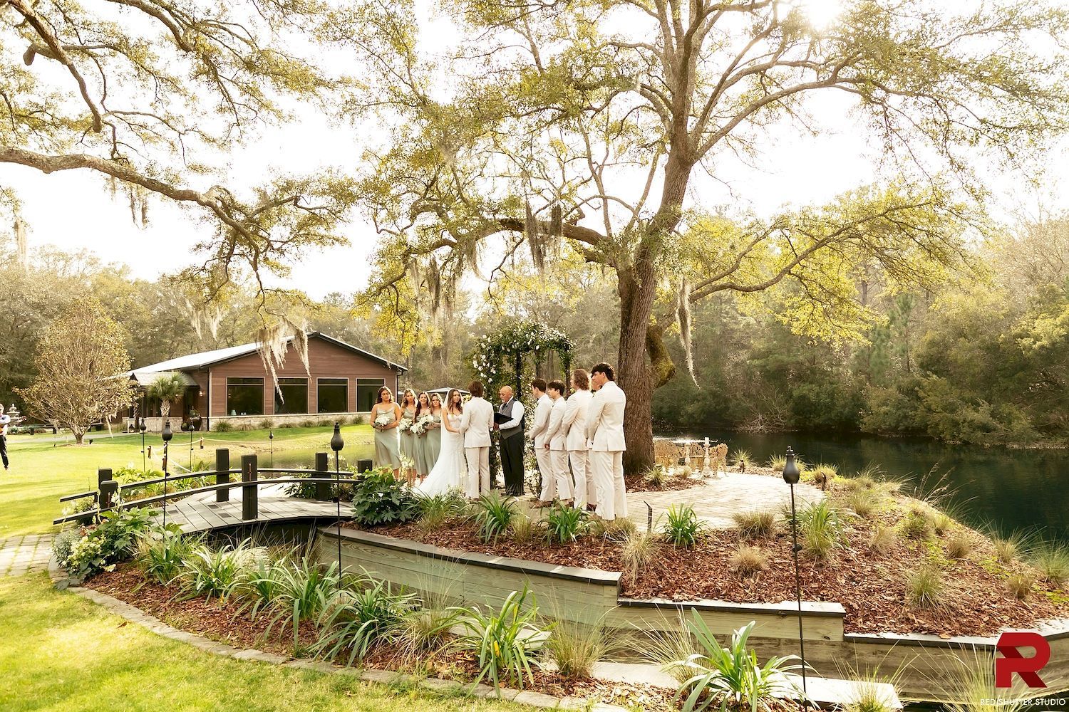Wedding party standing in a garden by a pond, with a gazebo and large trees in the background.