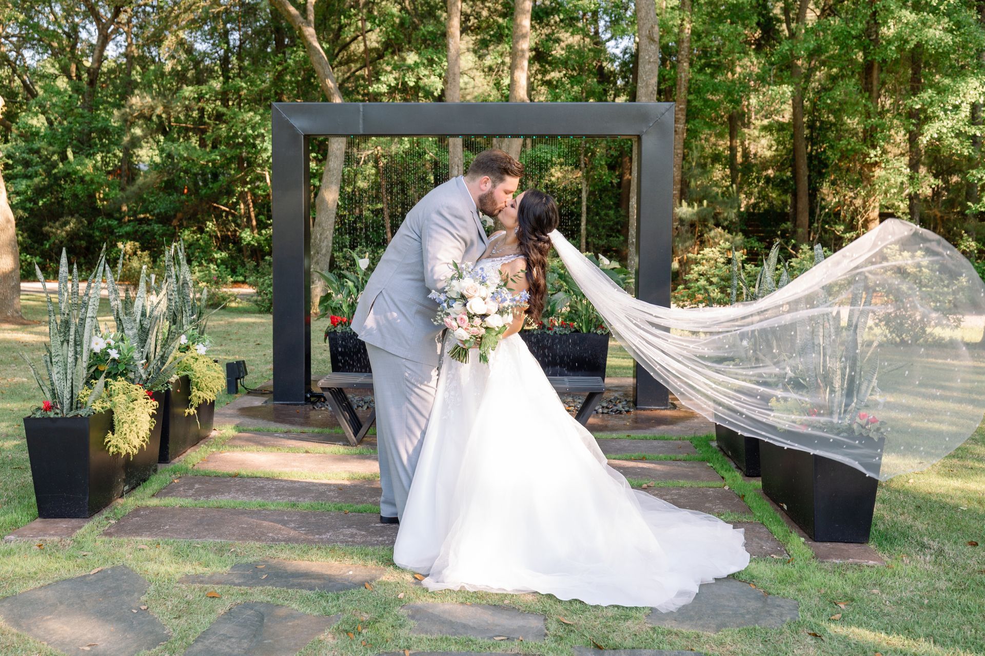 Bride and groom kissing in a garden, with the veil flowing behind her under a black archway.