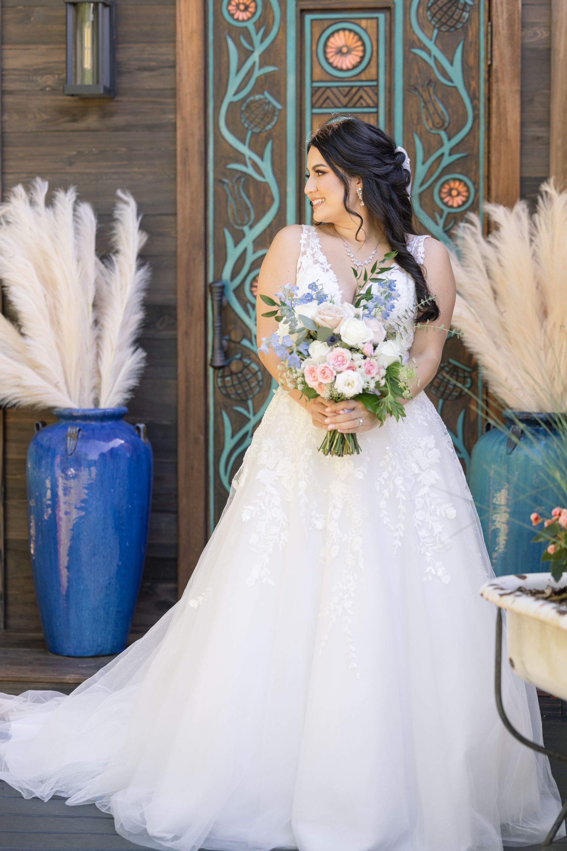Bride in white gown holding bouquet, standing before ornate door with blue vases and pampas grass