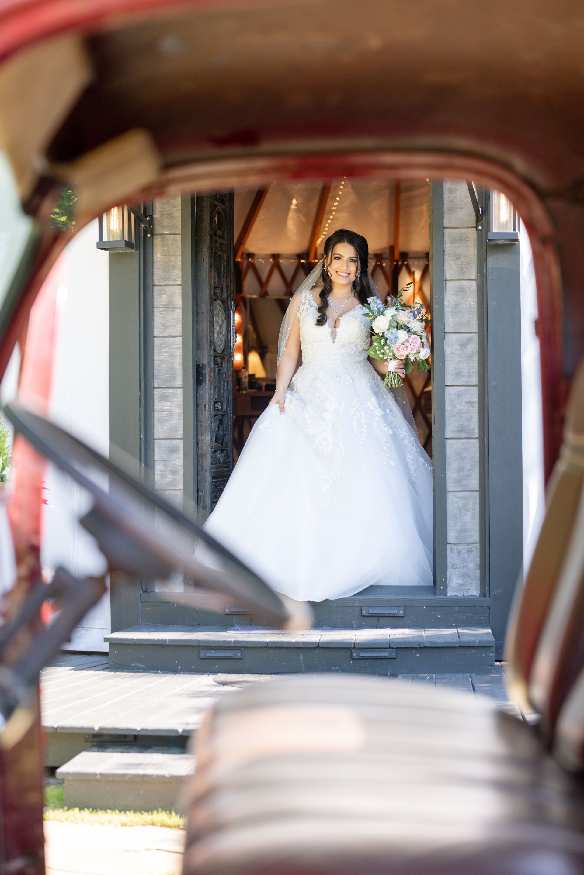 Bride in white gown holding bouquet, standing in a doorway framed by a vintage car interior