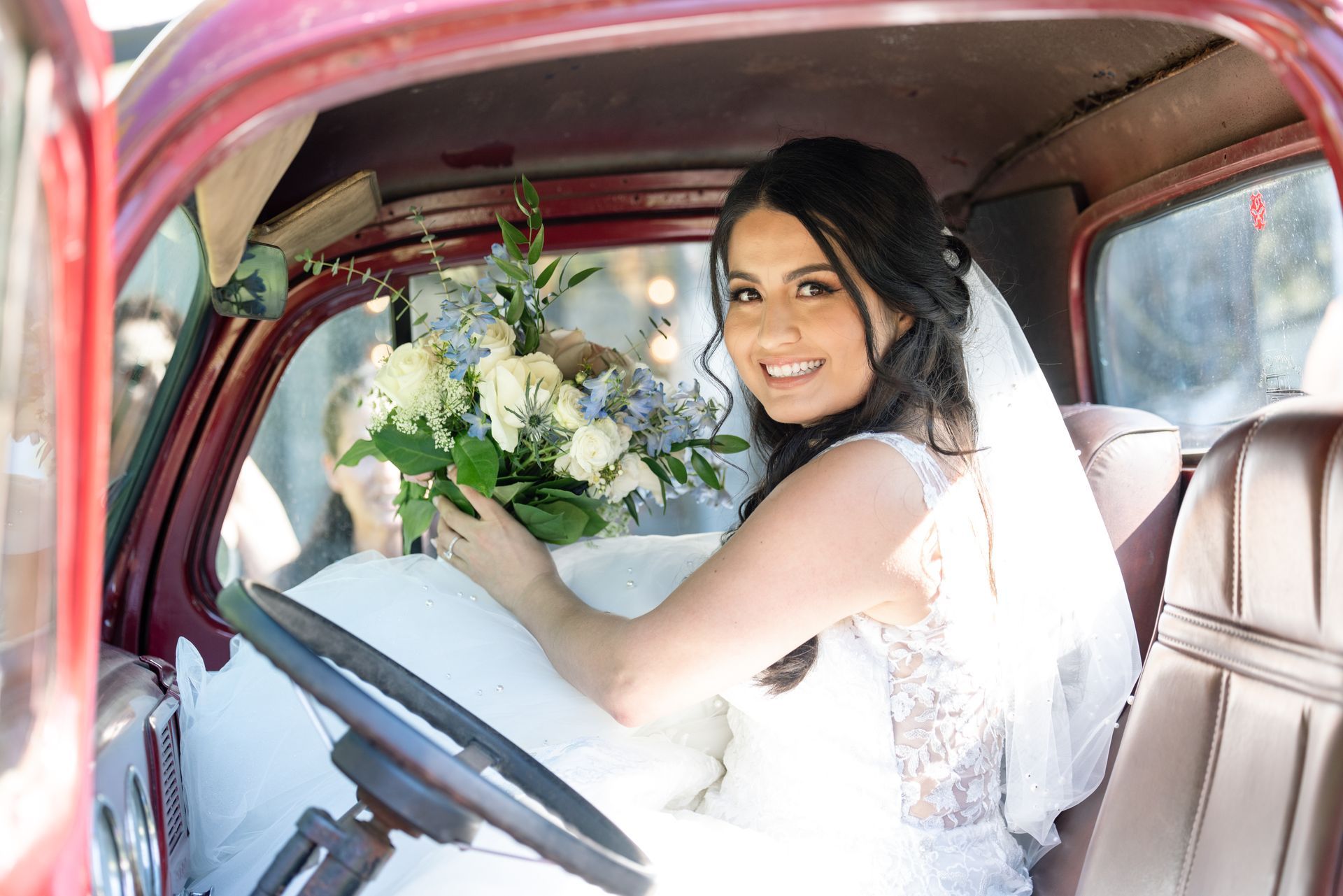 Bride in a white dress holding a bouquet, smiling from the driver’s seat of a vintage pink car