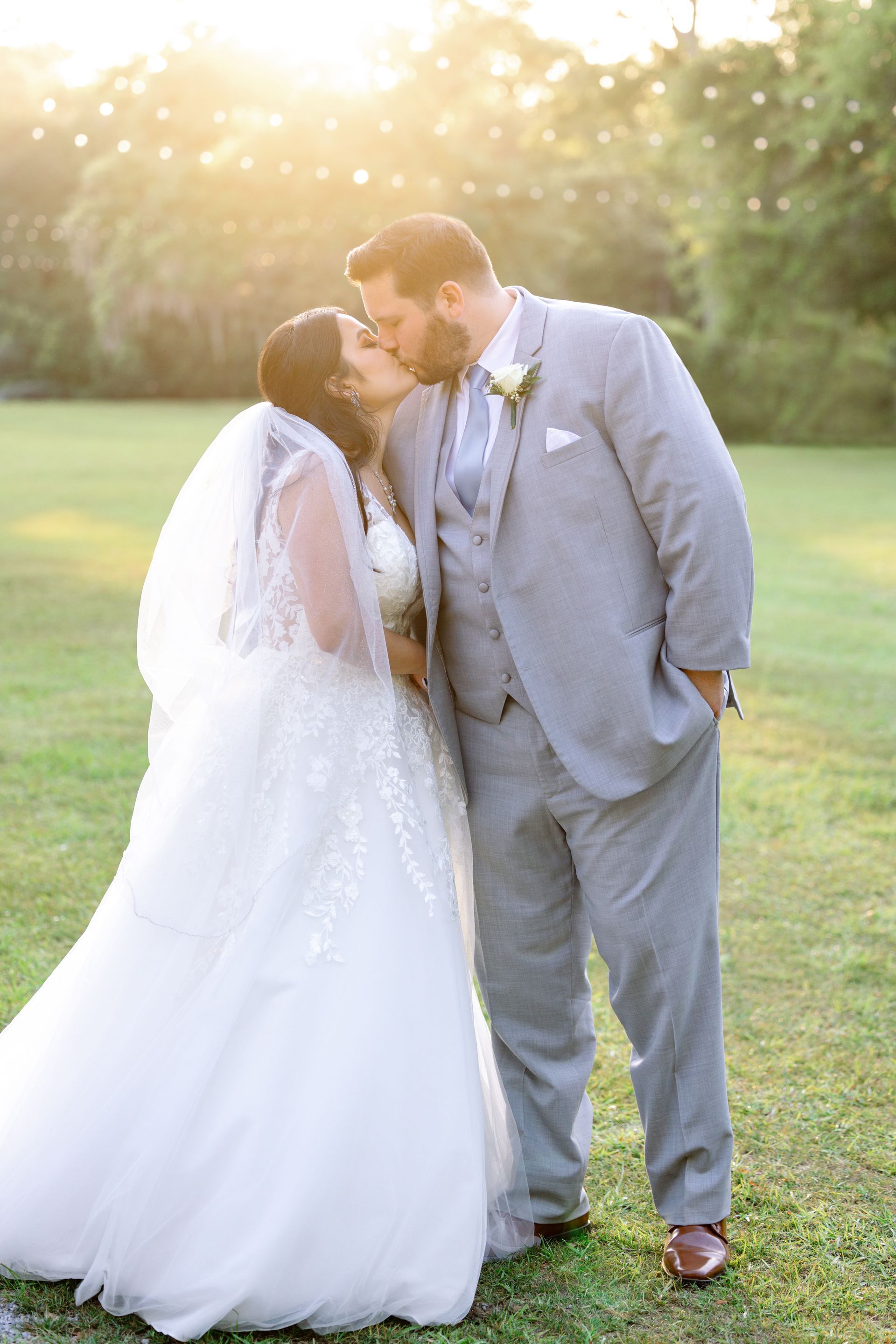 Bride and groom kiss in a sunlit outdoor field, with the bride in a white gown and the groom in a gray suit.