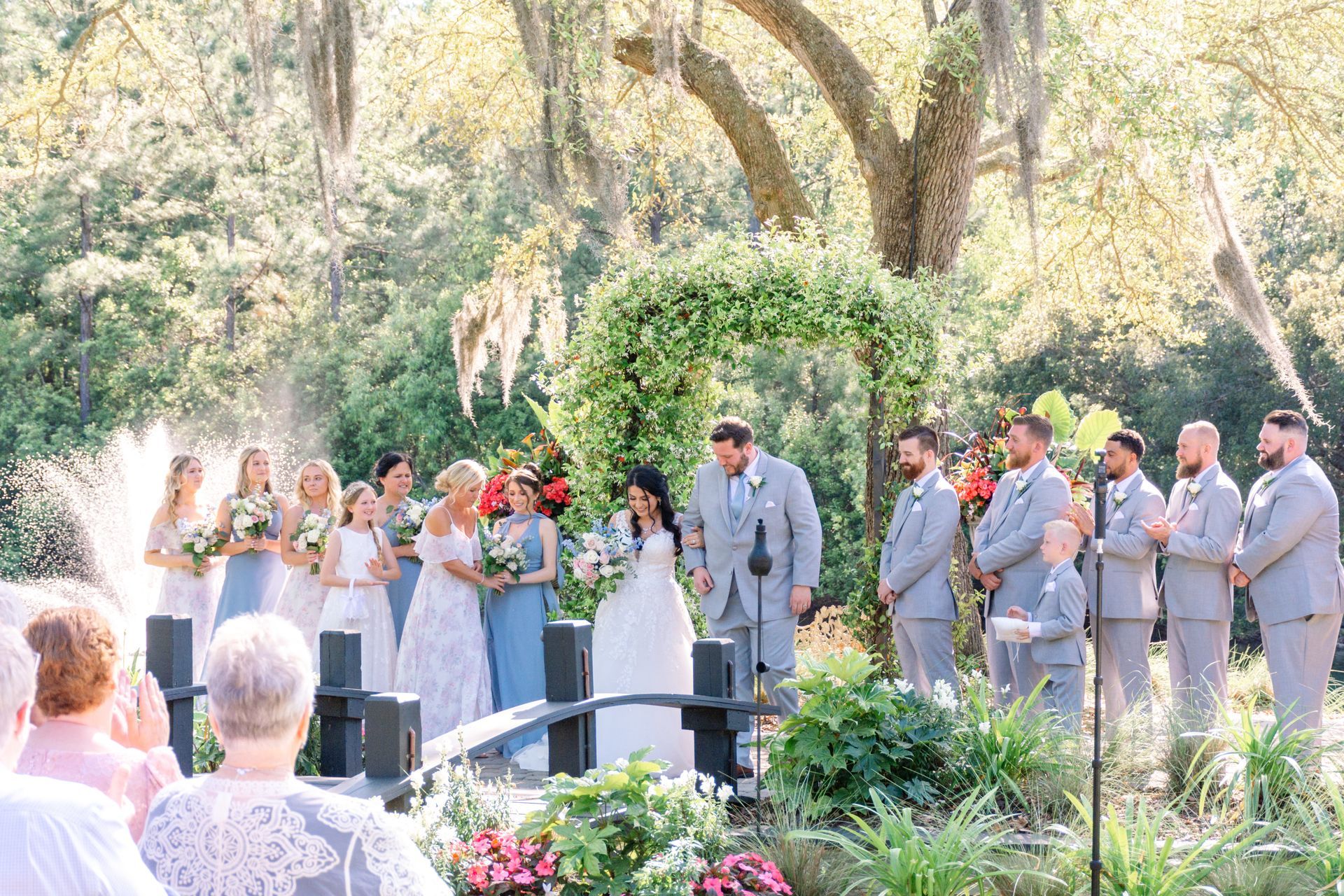 Outdoor wedding ceremony under trees, bride and groom with attendants standing near floral arrangements.