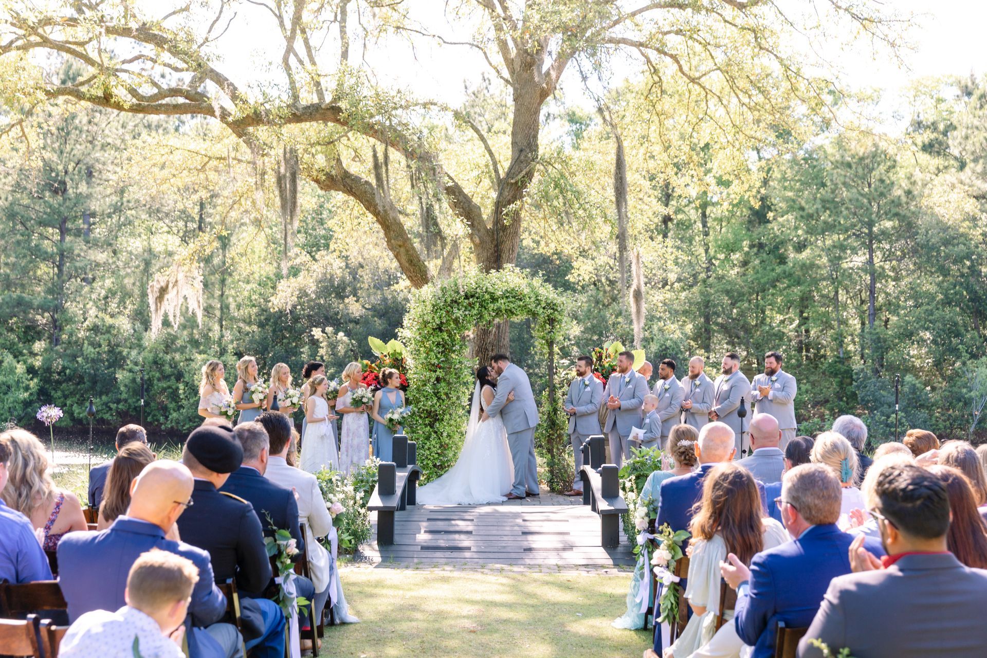 Outdoor wedding ceremony under a large tree, with guests seated facing an altar and officiant.