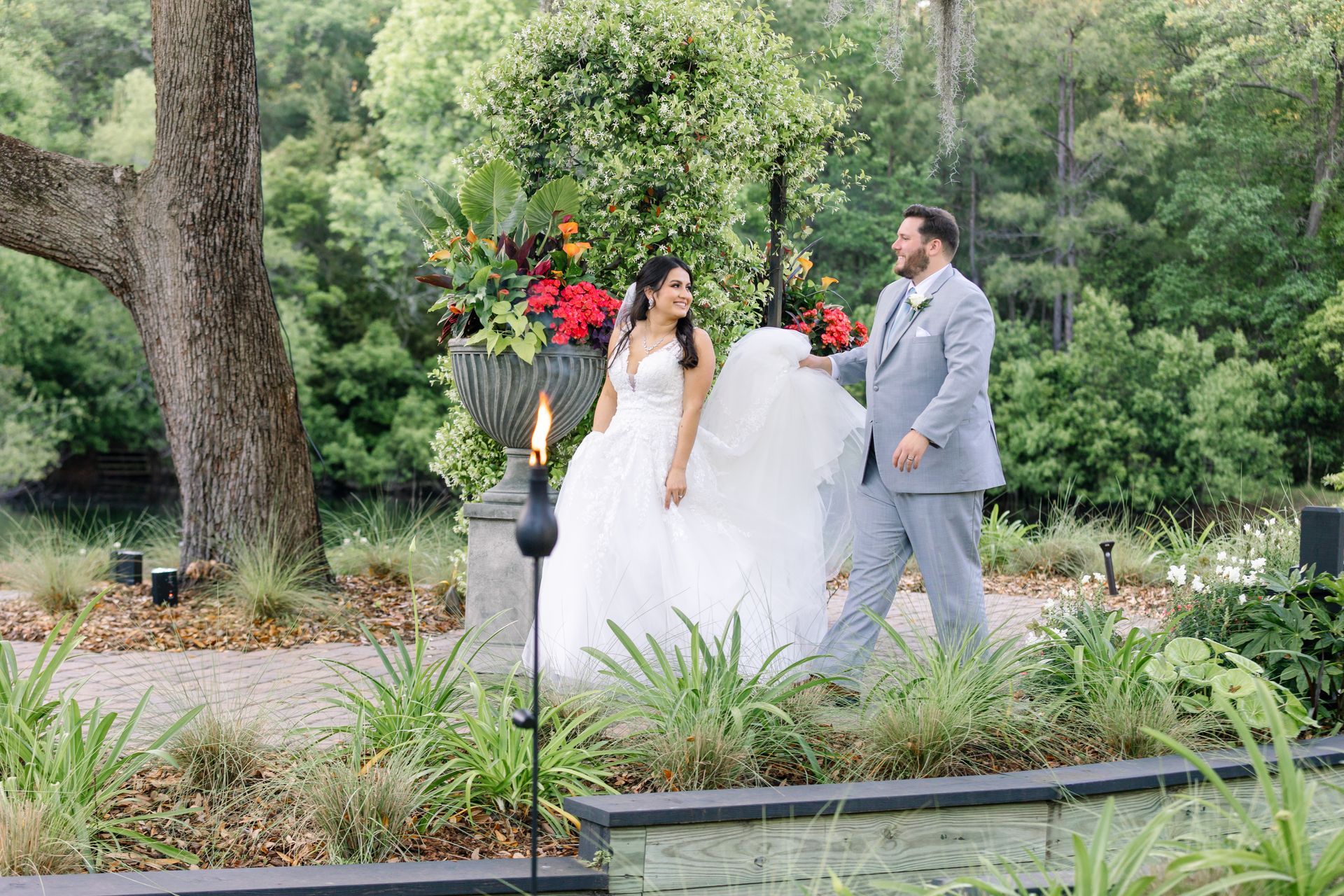 Bride and groom in white and gray walk through a lush garden wedding path with red floral arrangements