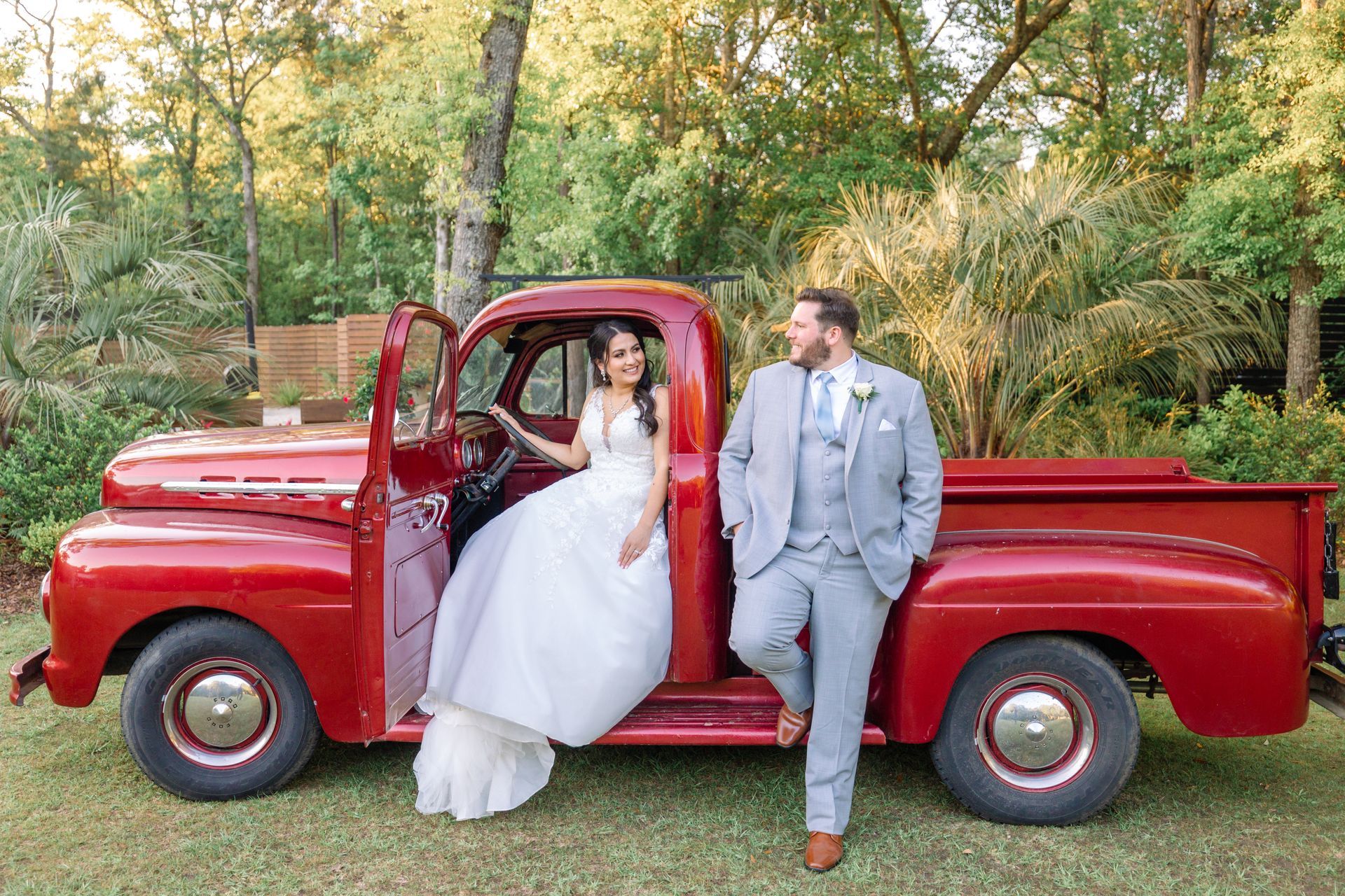 Bride and groom pose in a vintage red pickup truck outdoors in a wooded setting.