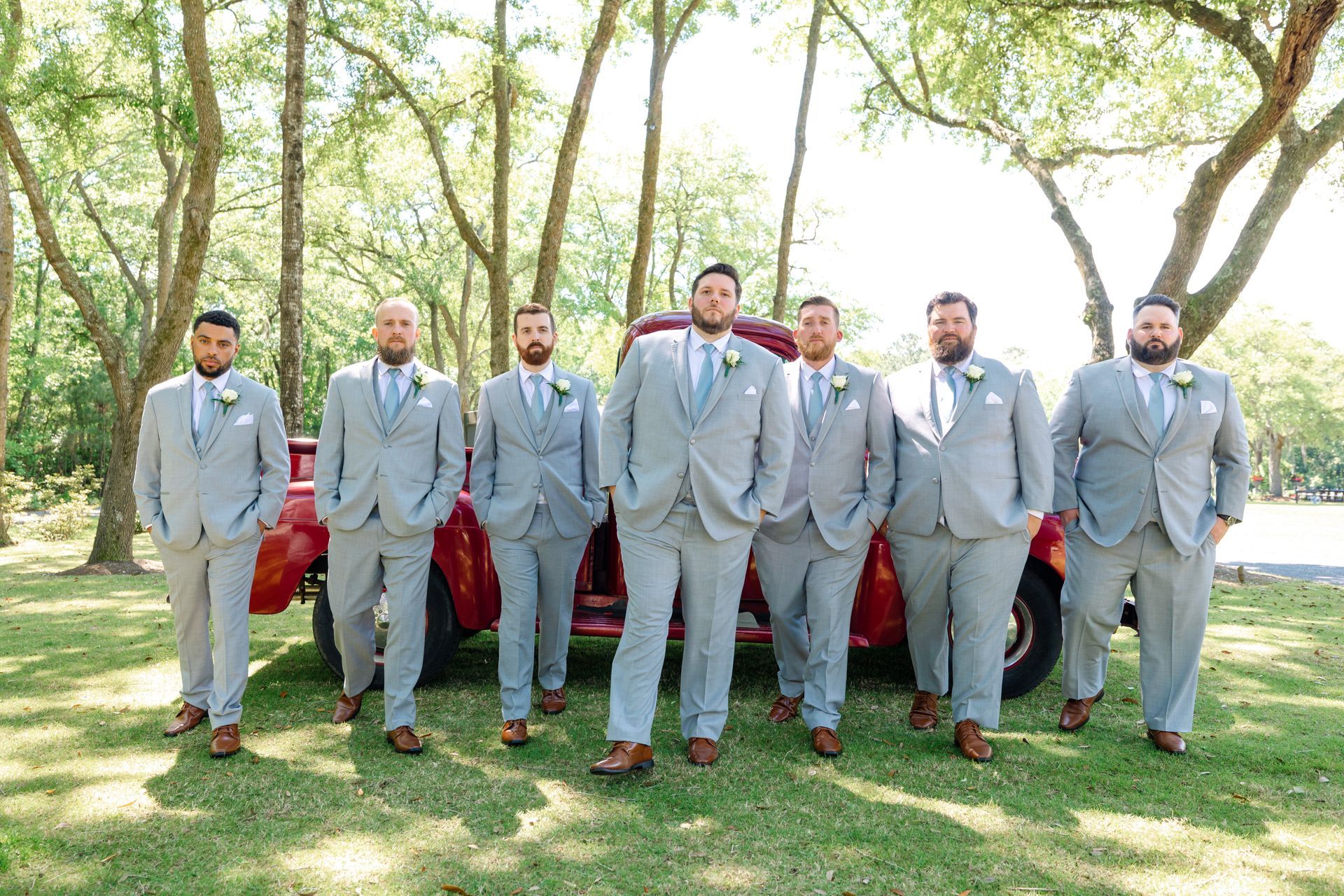 Seven people in gray suits posing outdoors in a sunny park, walking toward the camera.