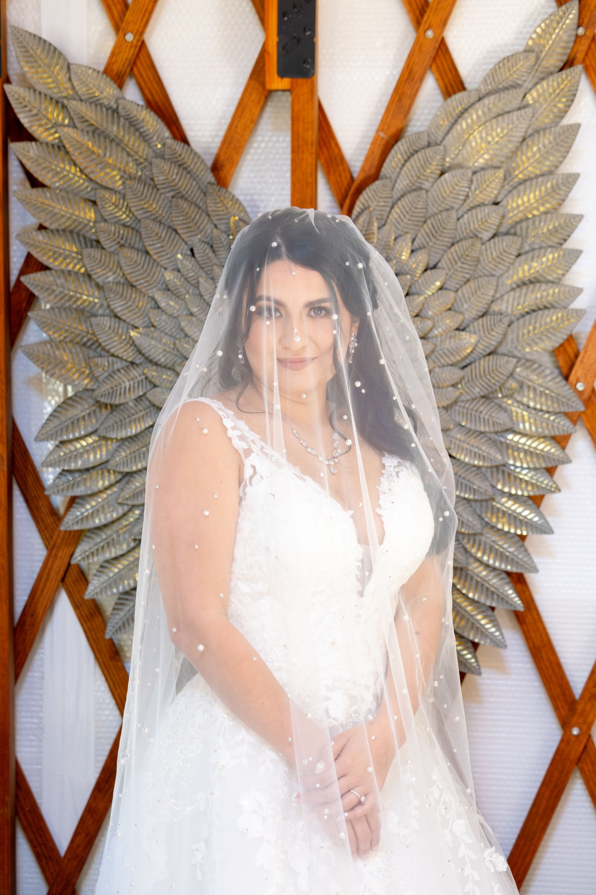 Woman in a white dress and veil posing in front of large metallic wings.