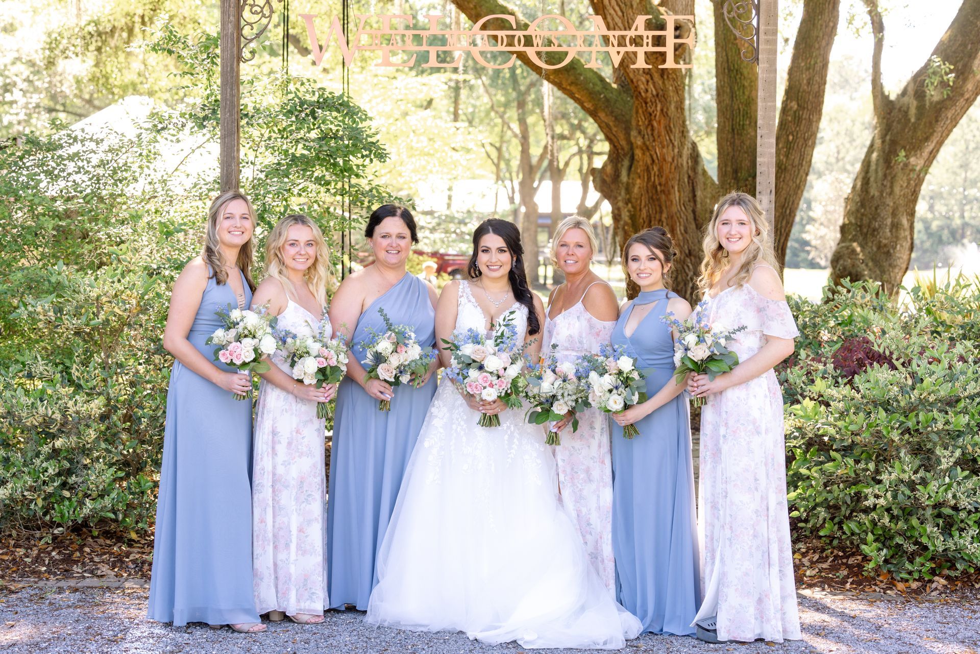Bridal party in lavender dresses and a white gown posing outdoors under trees