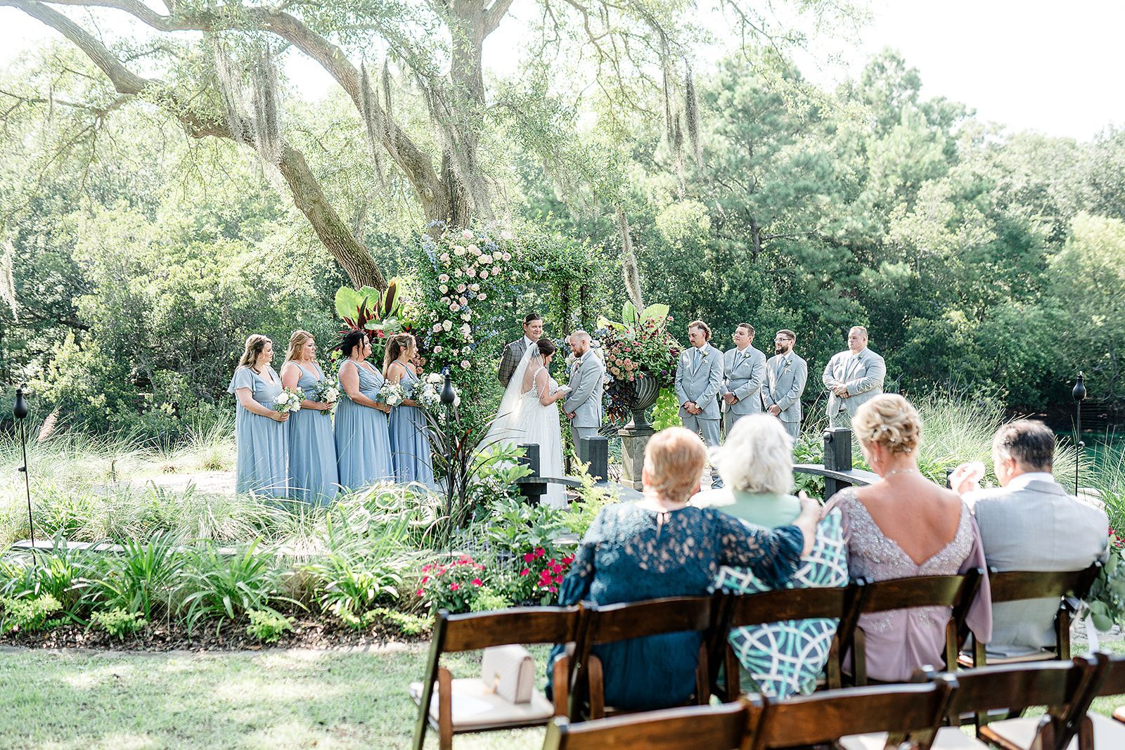Outdoor wedding ceremony in a garden, with bridal party standing before seated guests on benches.