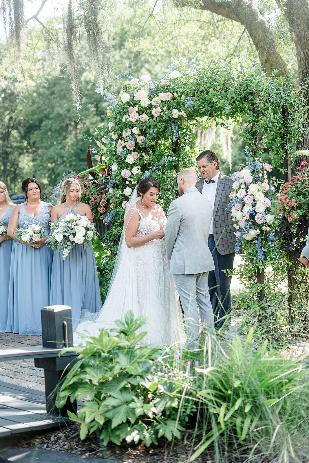 Wedding couple standing beneath a floral arch outdoors, with bridesmaids in blue dresses nearby.