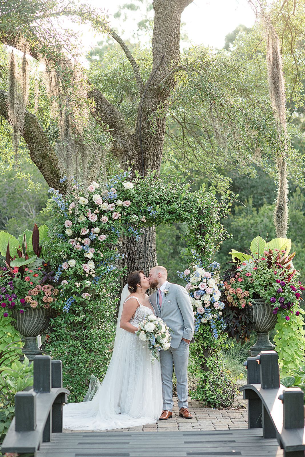 Bride and groom kiss beneath a flower-filled arch in an outdoor garden ceremony
