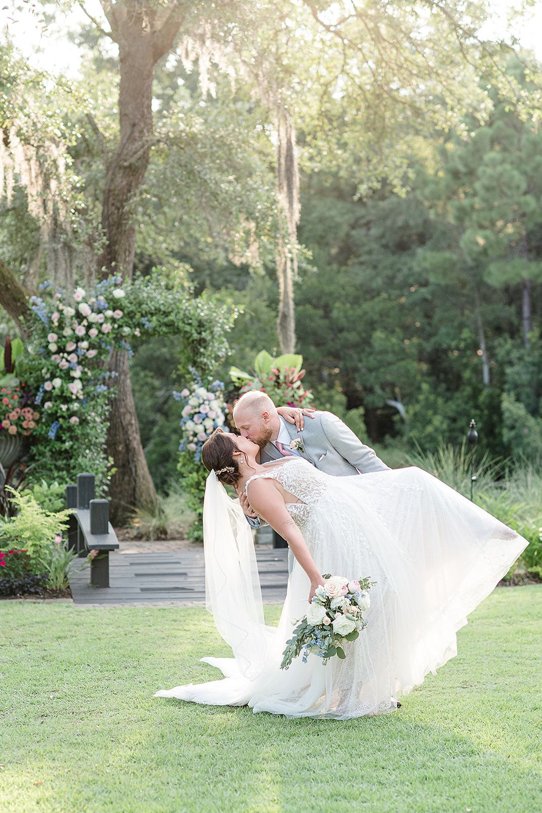 Bride and groom kiss in a garden as her white dress and veil billow in the breeze.