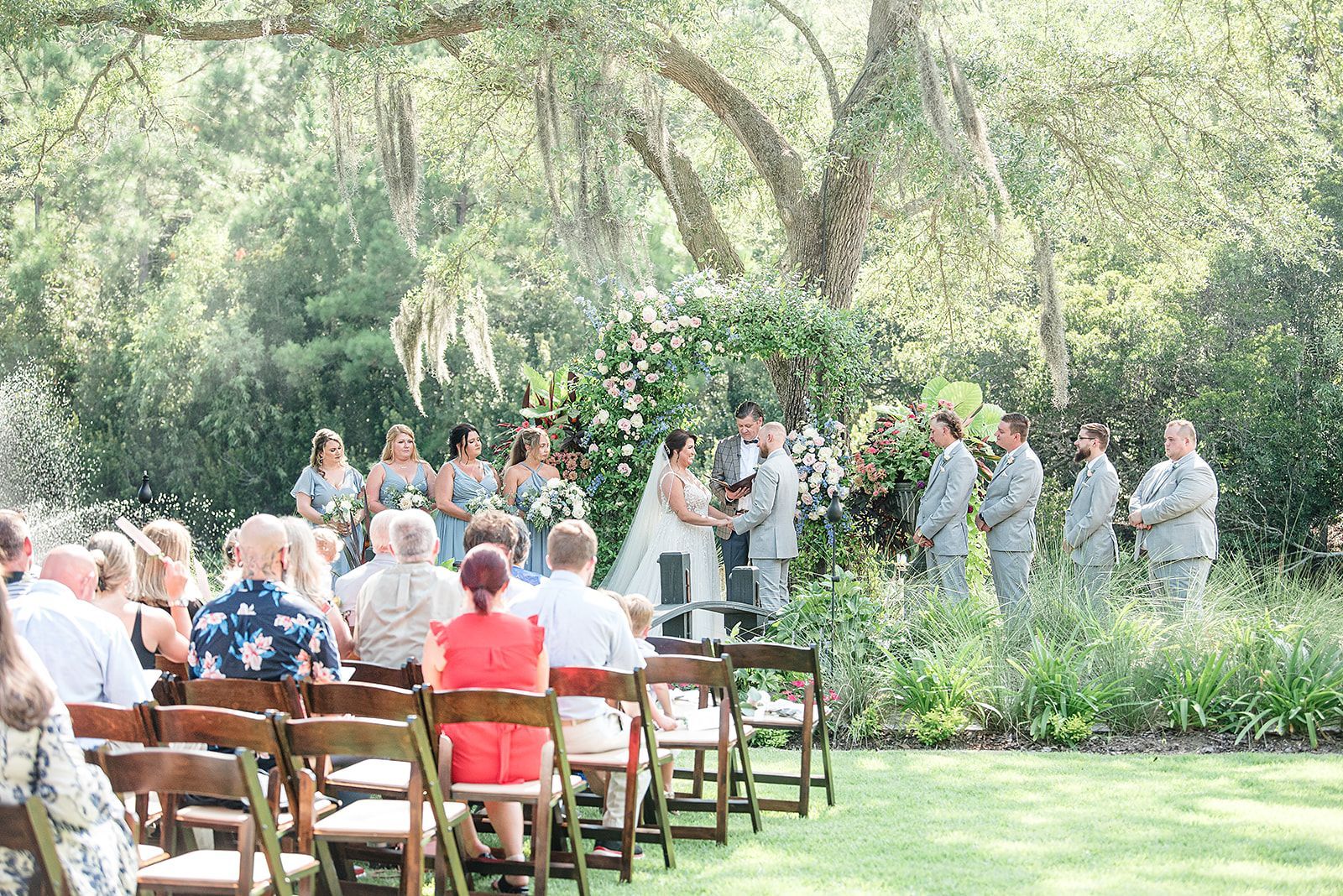 Outdoor wedding ceremony under large trees with guests seated on wooden benches and people gathered at the altar.