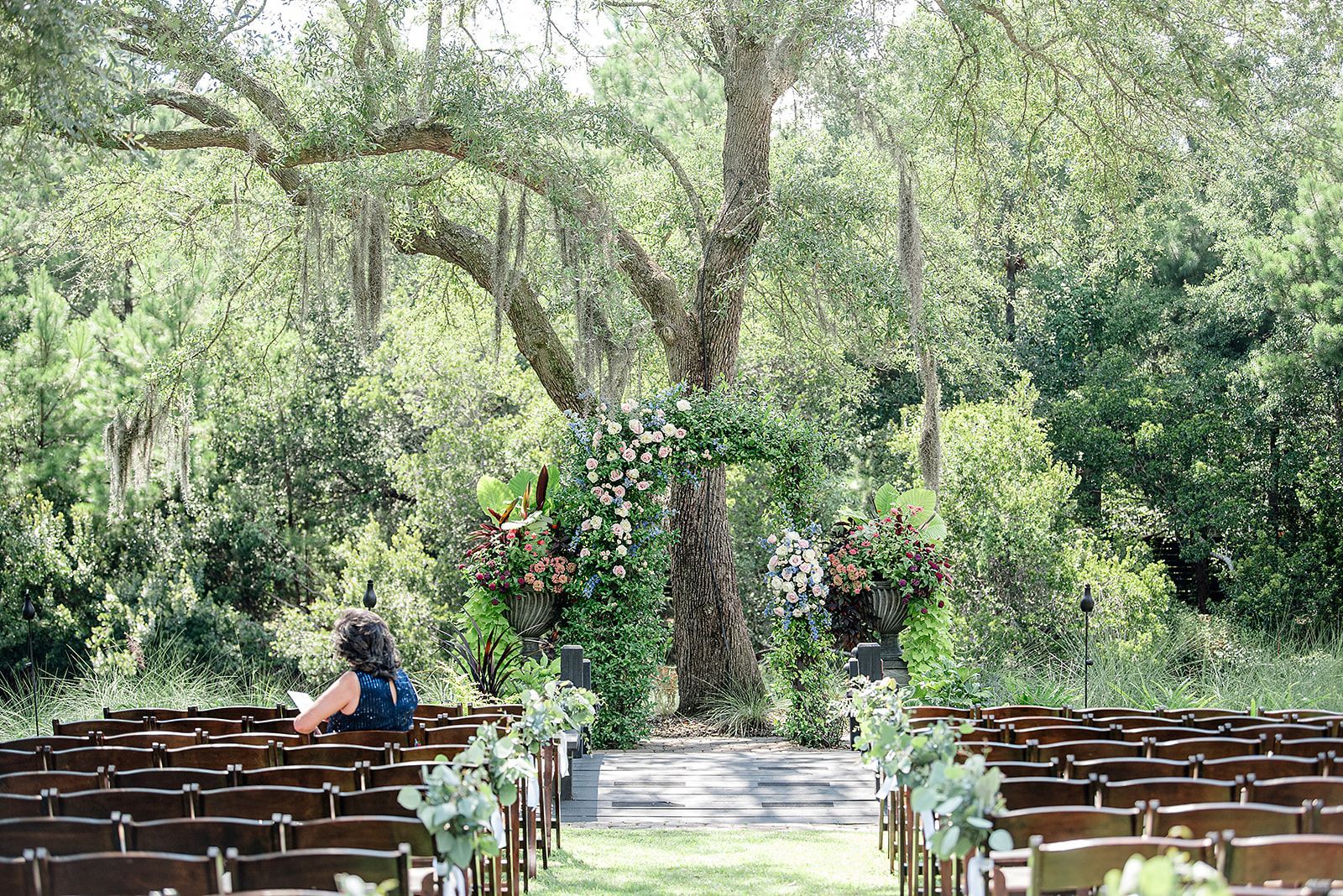 Outdoor wedding aisle beneath trees, with white chairs and a floral arch in the center.