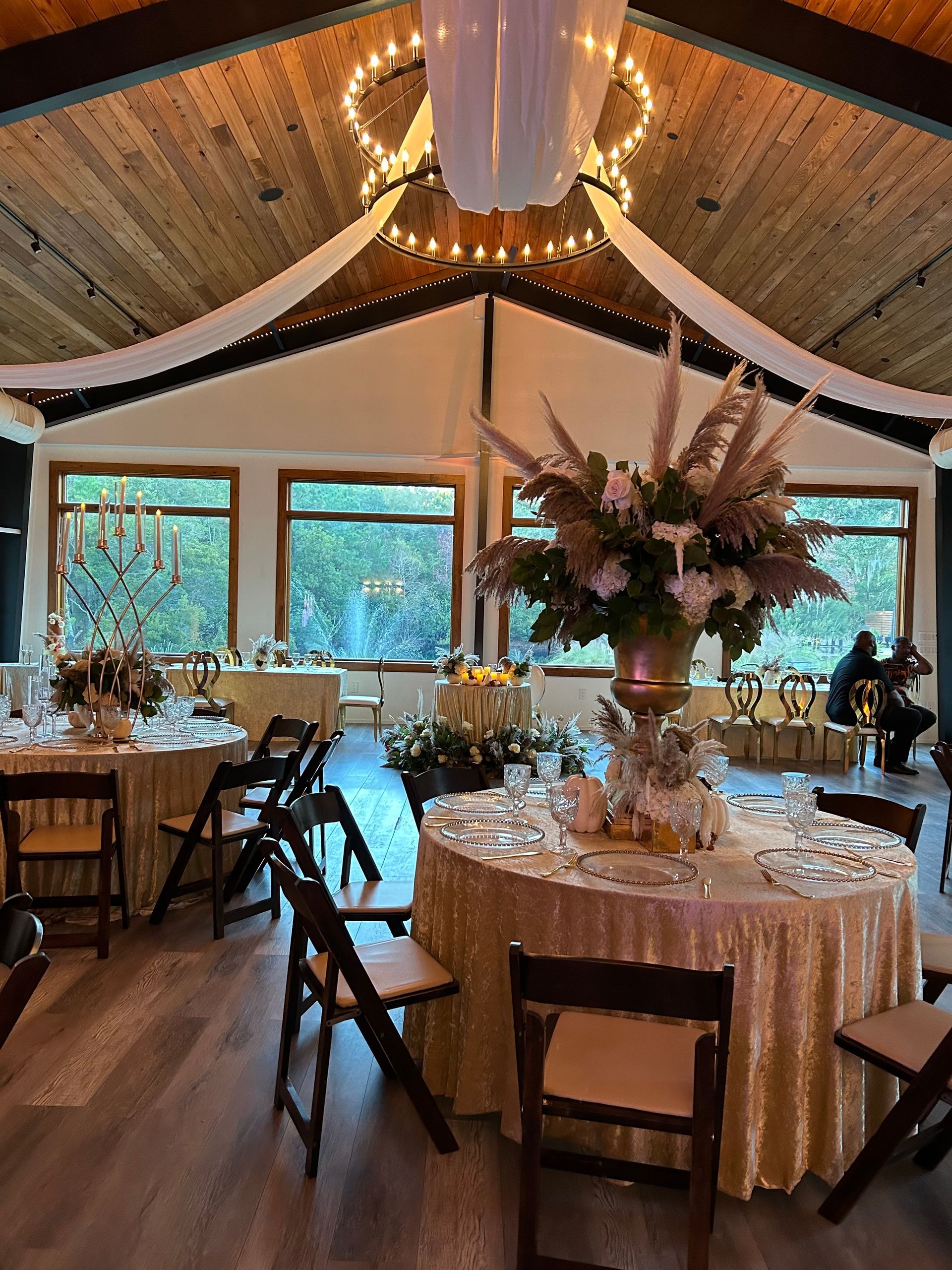 Elegant reception hall with round tables, floral centerpieces, and warm string lights under a high wooden ceiling
