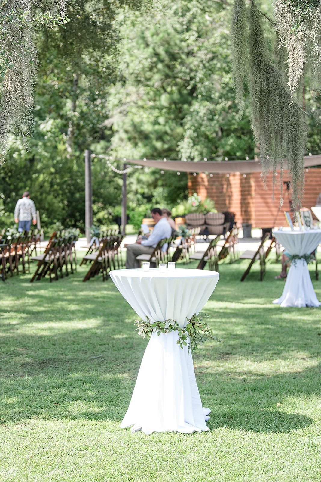 Outdoor garden reception with round cocktail tables and rows of chairs on a grassy lawn under trees.