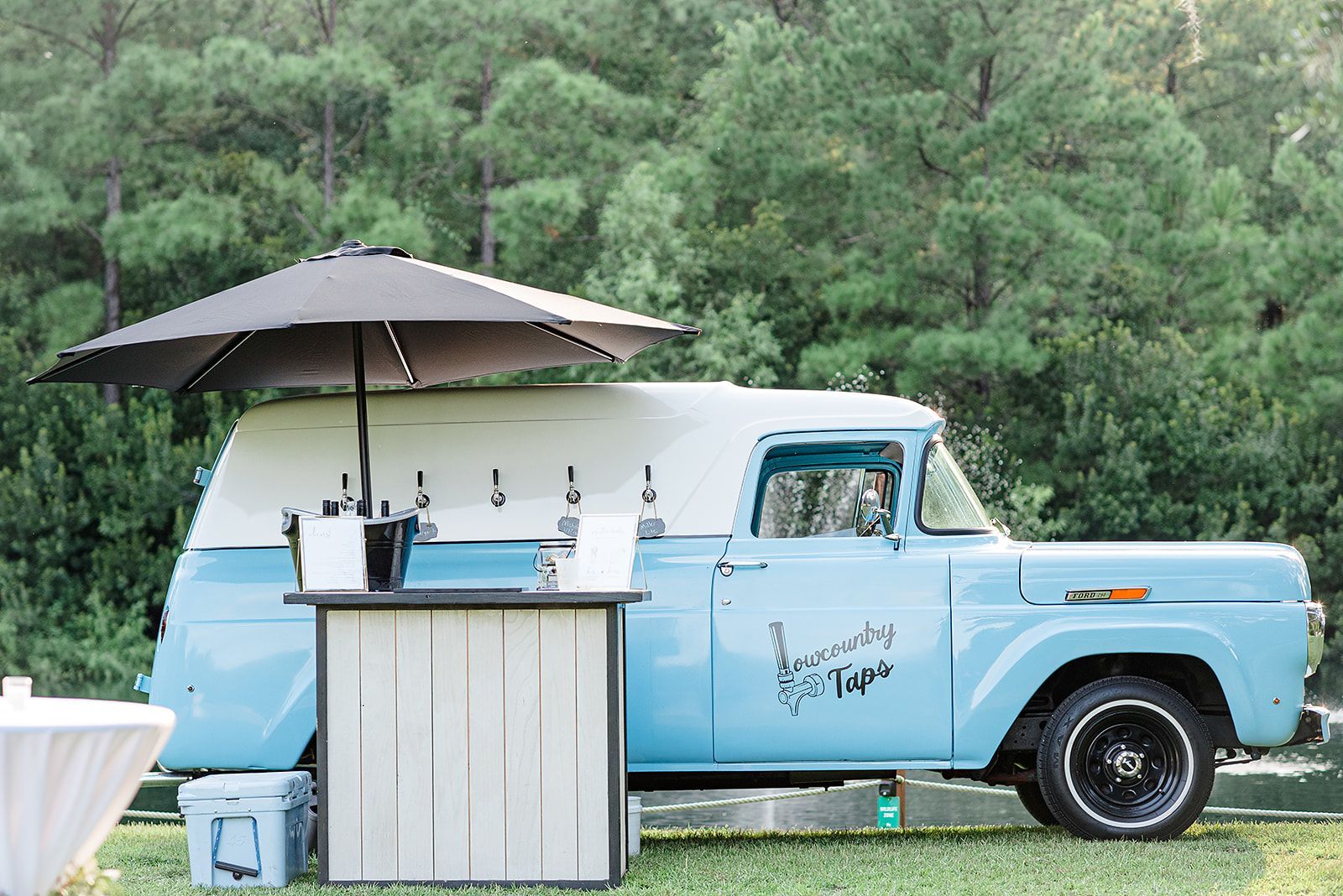 Light blue vintage truck beside a small bar under an umbrella in a grassy outdoor setting