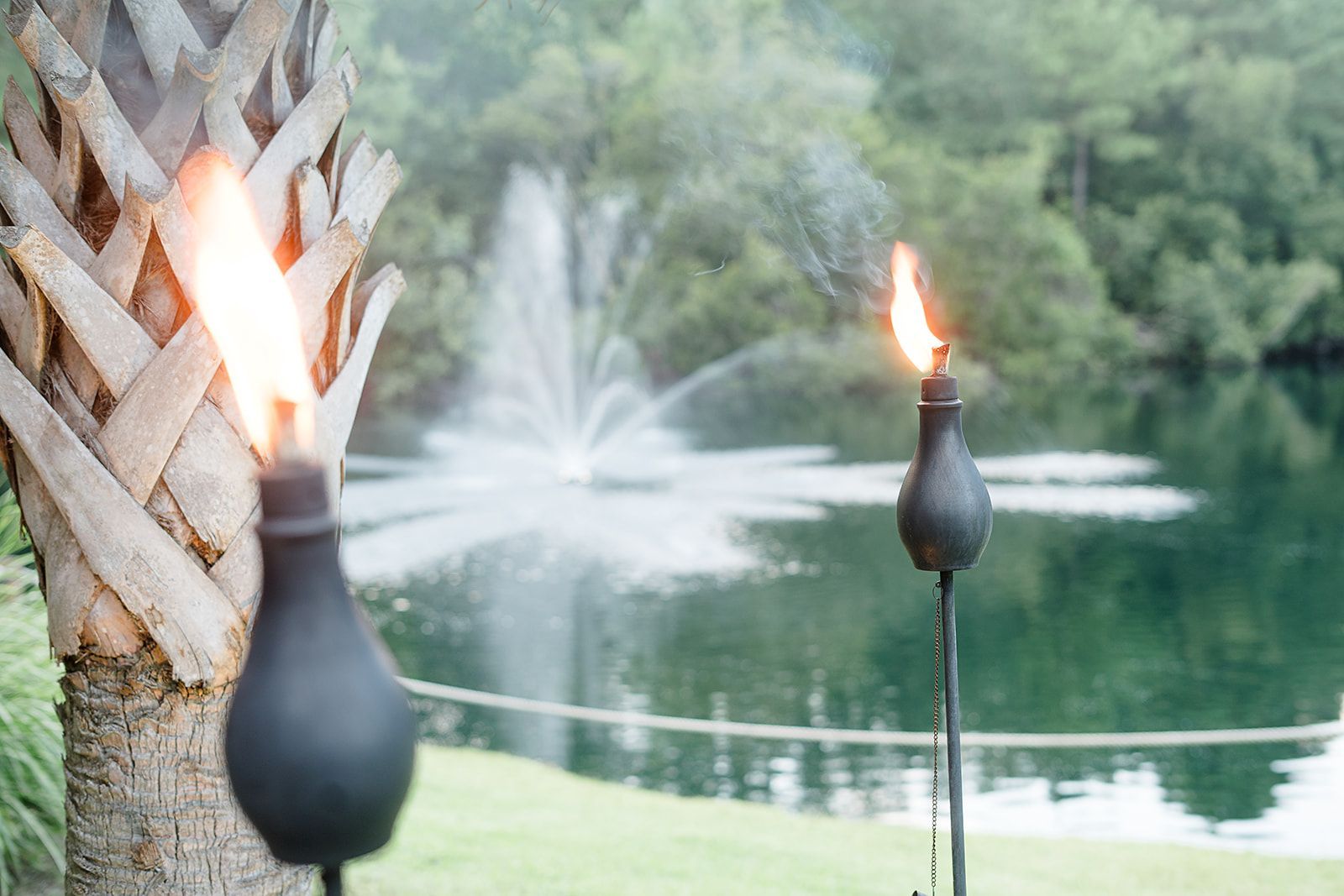 Two lit tiki torches by a pond with a fountain and trees in the background