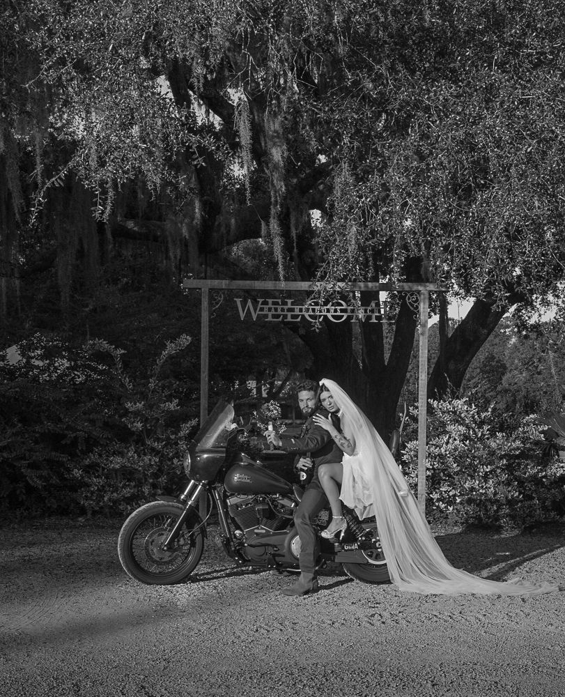 Bride and groom on a motorcycle under trees at night, with a long veil trailing behind them