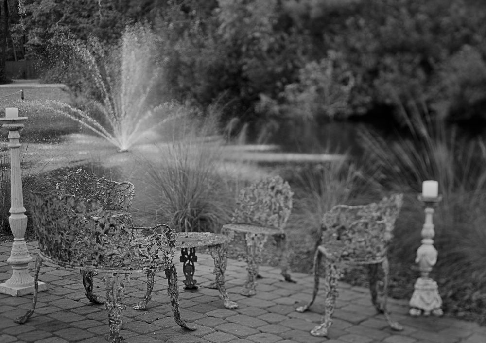 Garden chairs on a patio being sprayed by a sprinkler under trees.