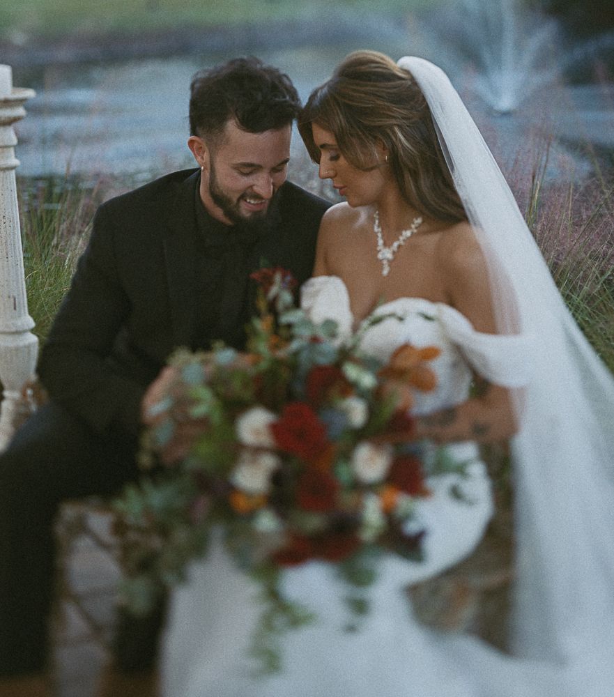 Bride and groom in white and black wedding attire, smiling together with a bouquet outdoors