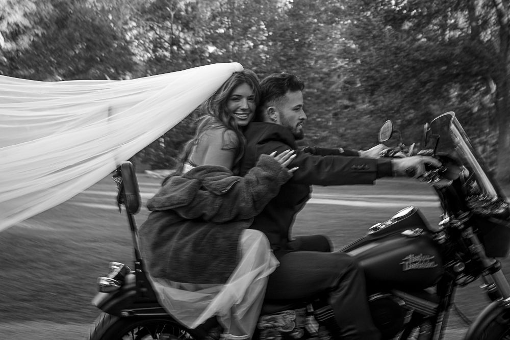 Two people riding a motorcycle, smiling under a billowing white sheet on a road.