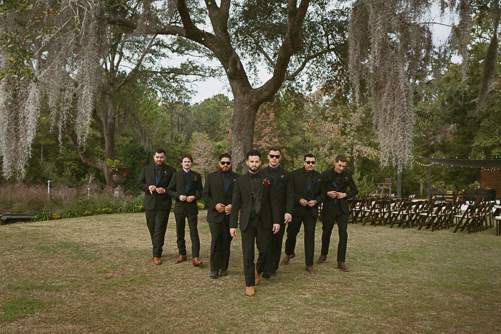 Group of seven people in black outfits walking toward the camera in a grassy park under a large tree.