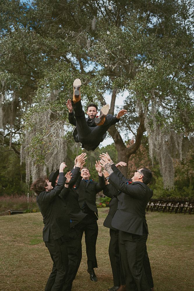 Group in dark clothing tossing a person into the air outdoors under trees.