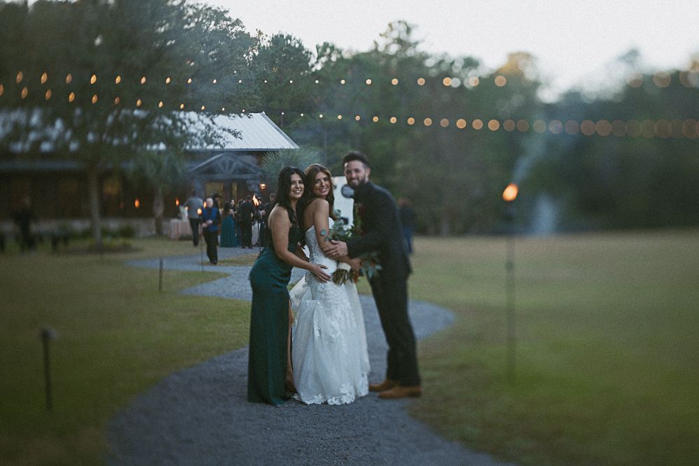 Three people posing outdoors at dusk, with string lights and a grassy field in the background.