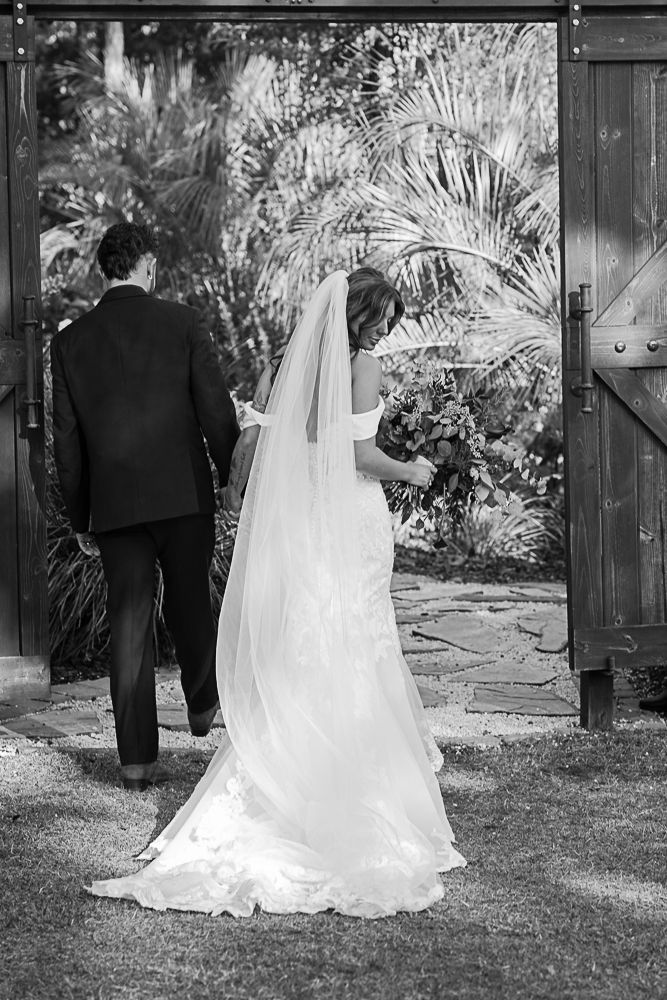 Bride and groom in formalwear walking through a garden gate, seen from behind.