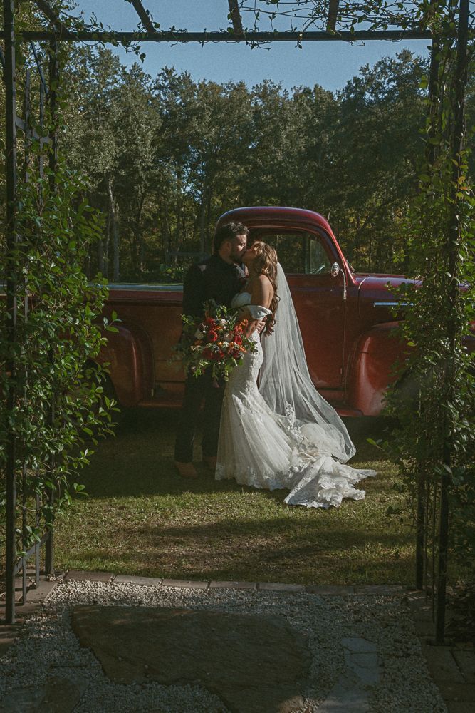 Bride in a white lace gown stands by a red pickup truck in a garden, holding a bouquet.