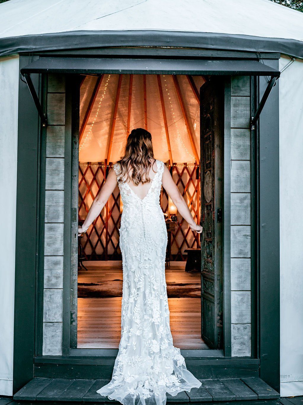 Woman in a white lace gown standing in a doorway with warm curtains behind her