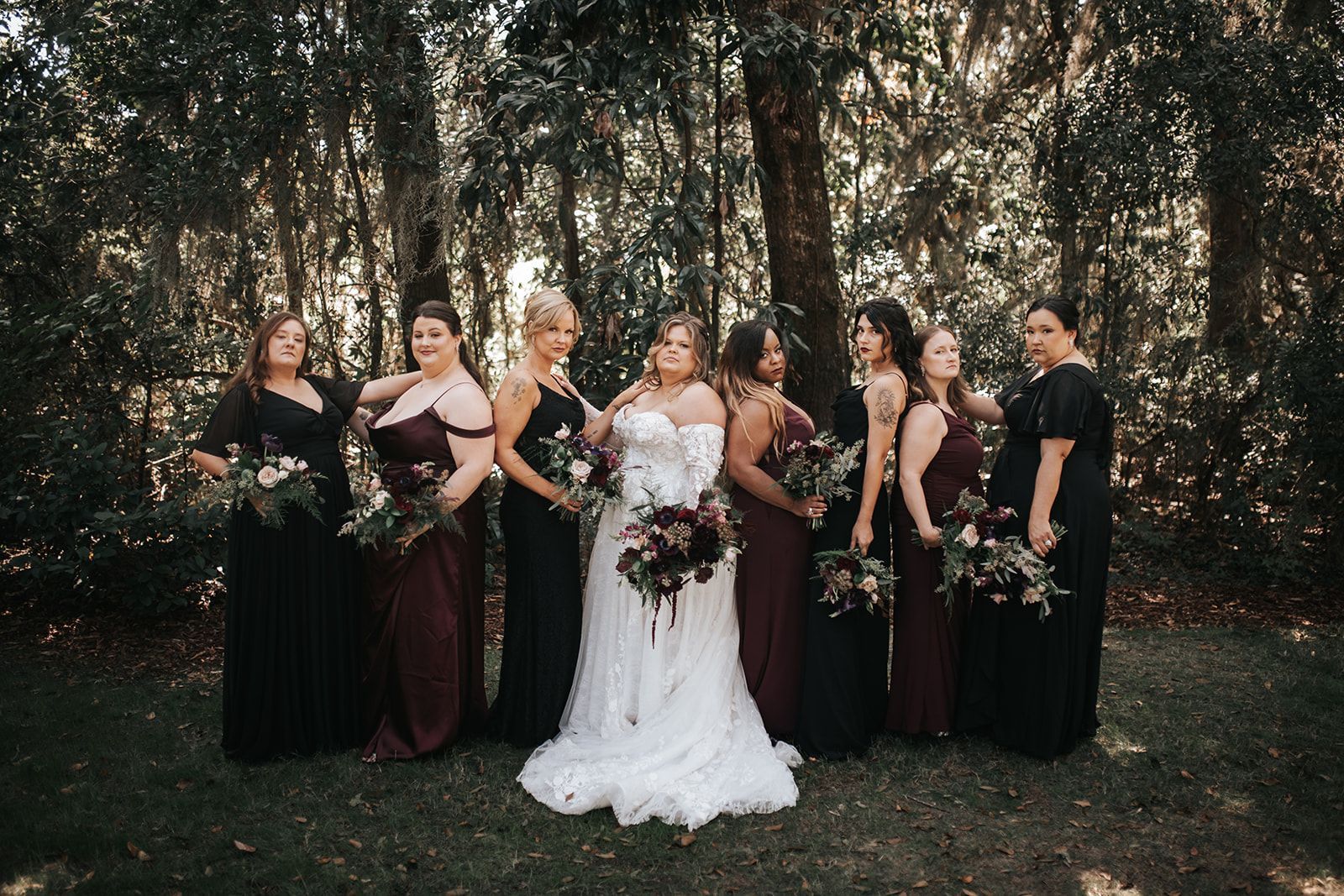 Group of six people in formal dresses holding bouquets in a wooded outdoor setting