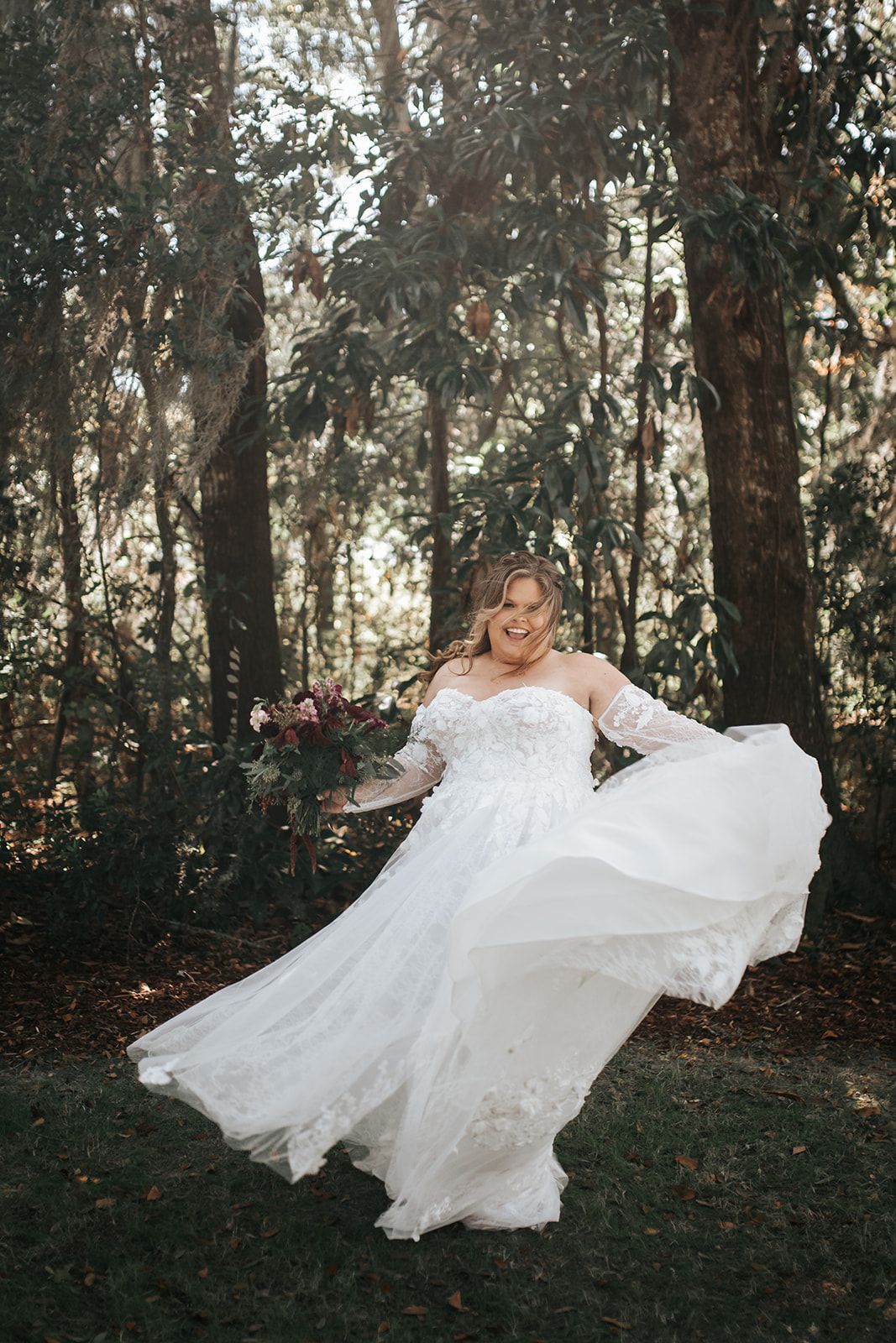 Bride in a flowing white wedding gown twirling in a wooded outdoor setting, smiling.
