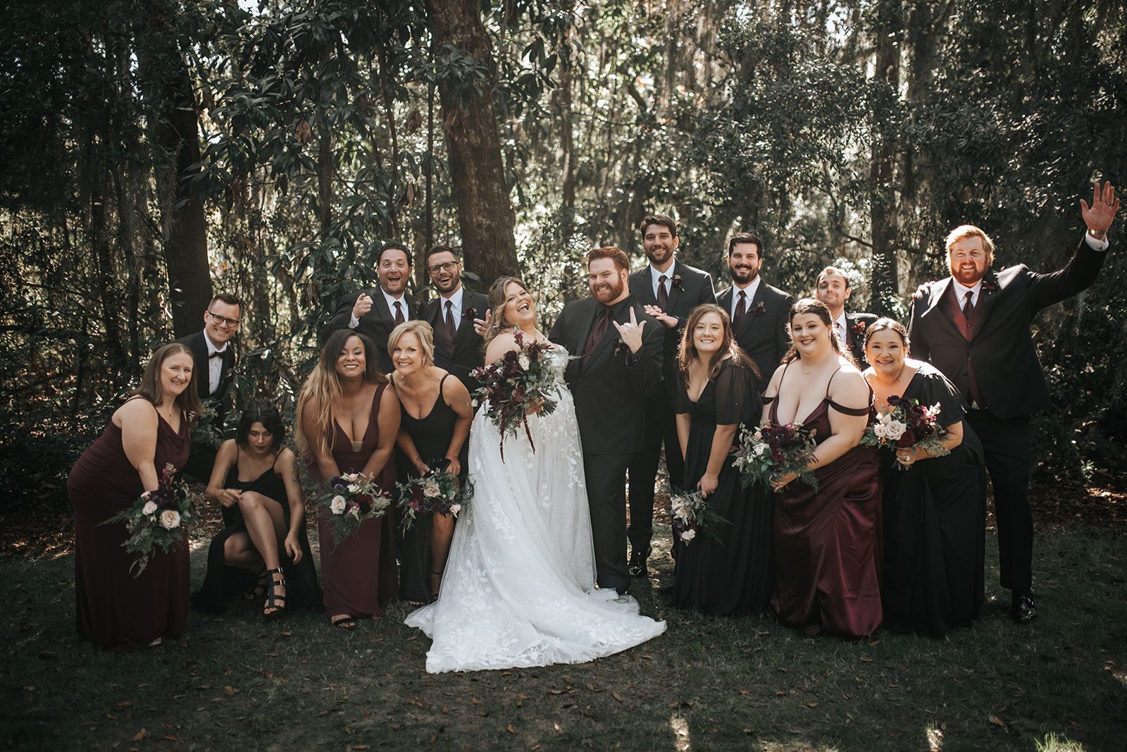 Wedding party posing outdoors in formal attire among trees, with bride in white gown at center