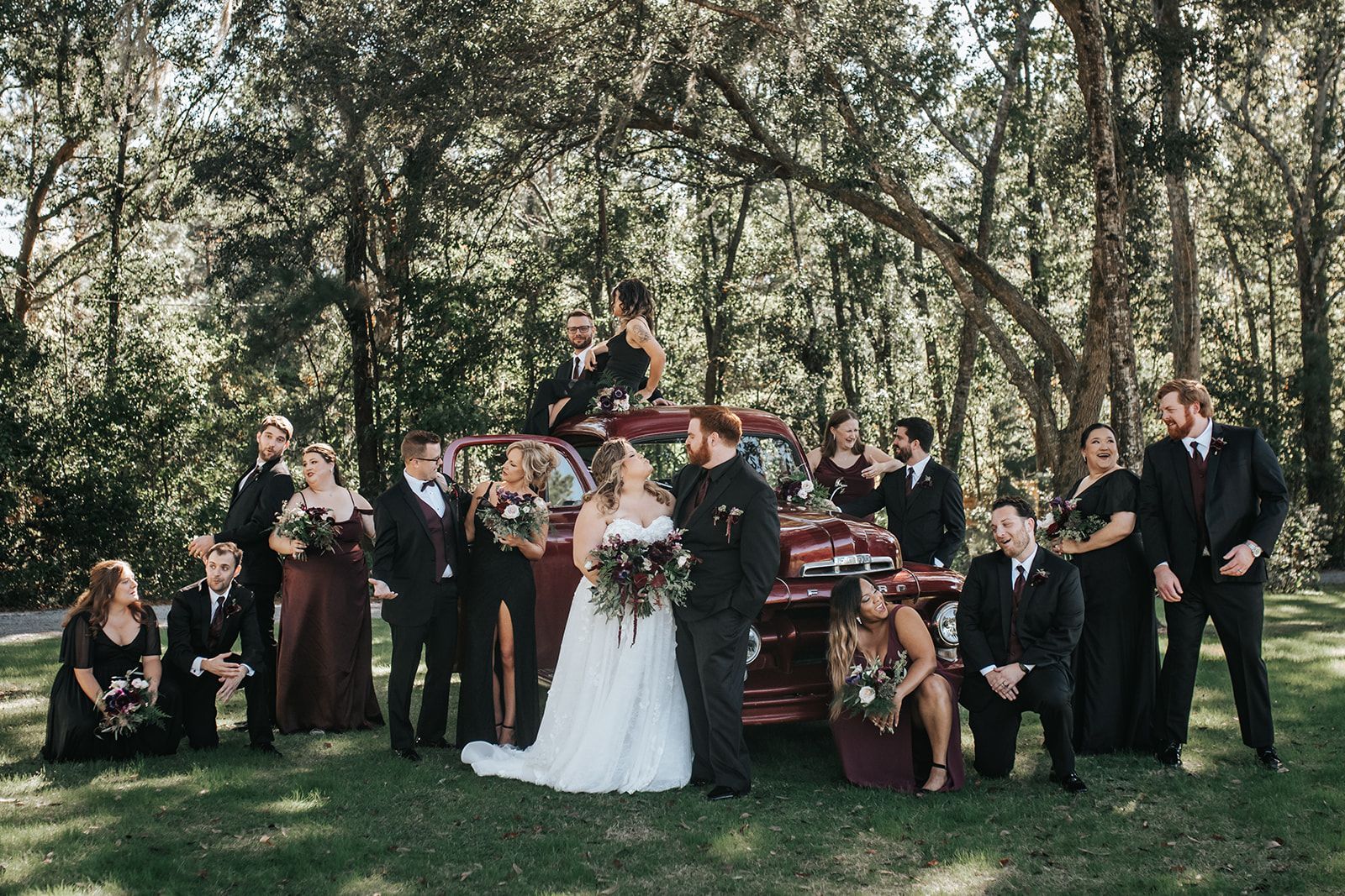 Wedding party posing outdoors among trees, with bride and groom centered and guests arranged around them.