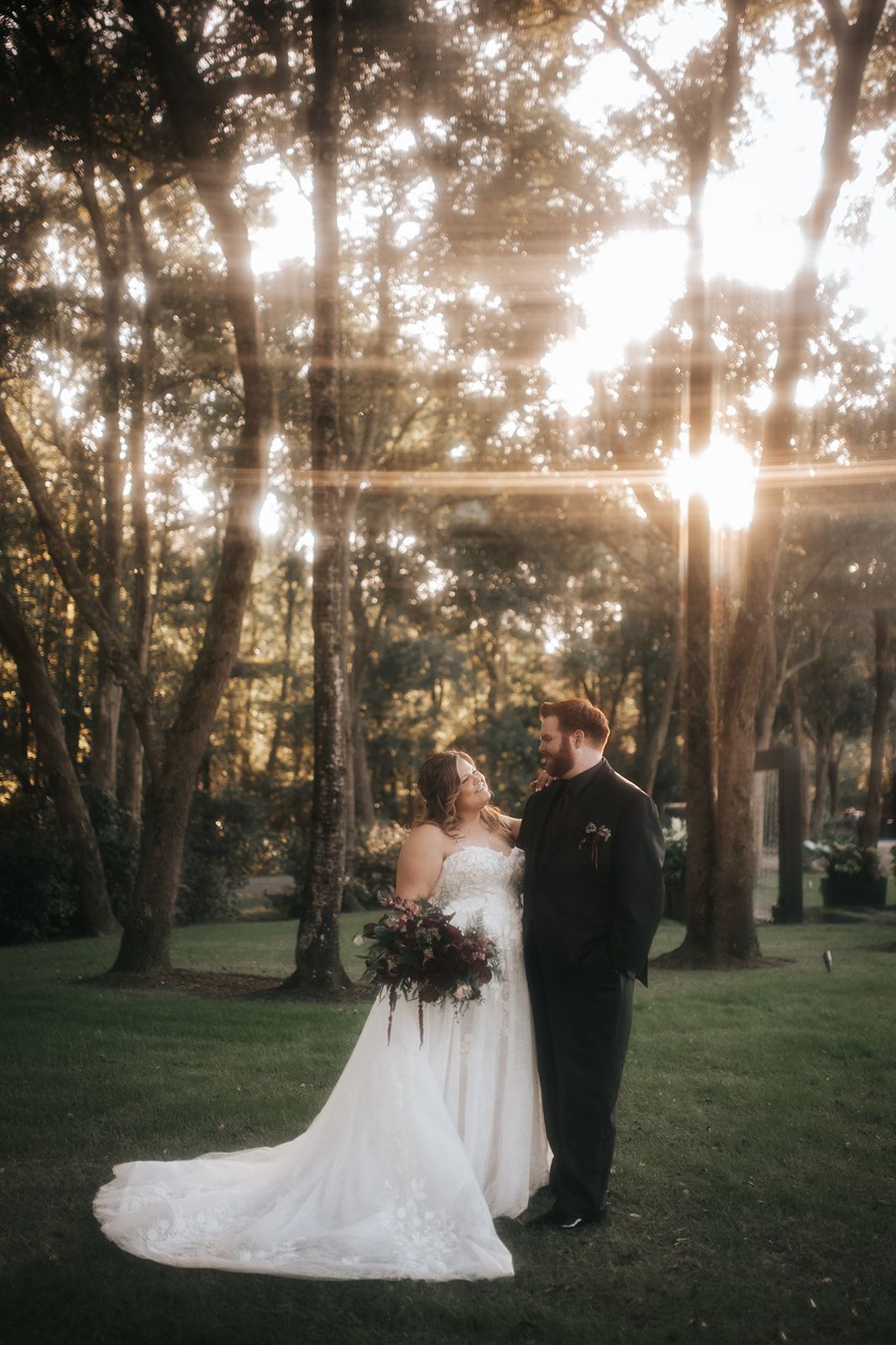 Bride and groom standing together in a sunlit wooded park, bride in a white gown holding a bouquet