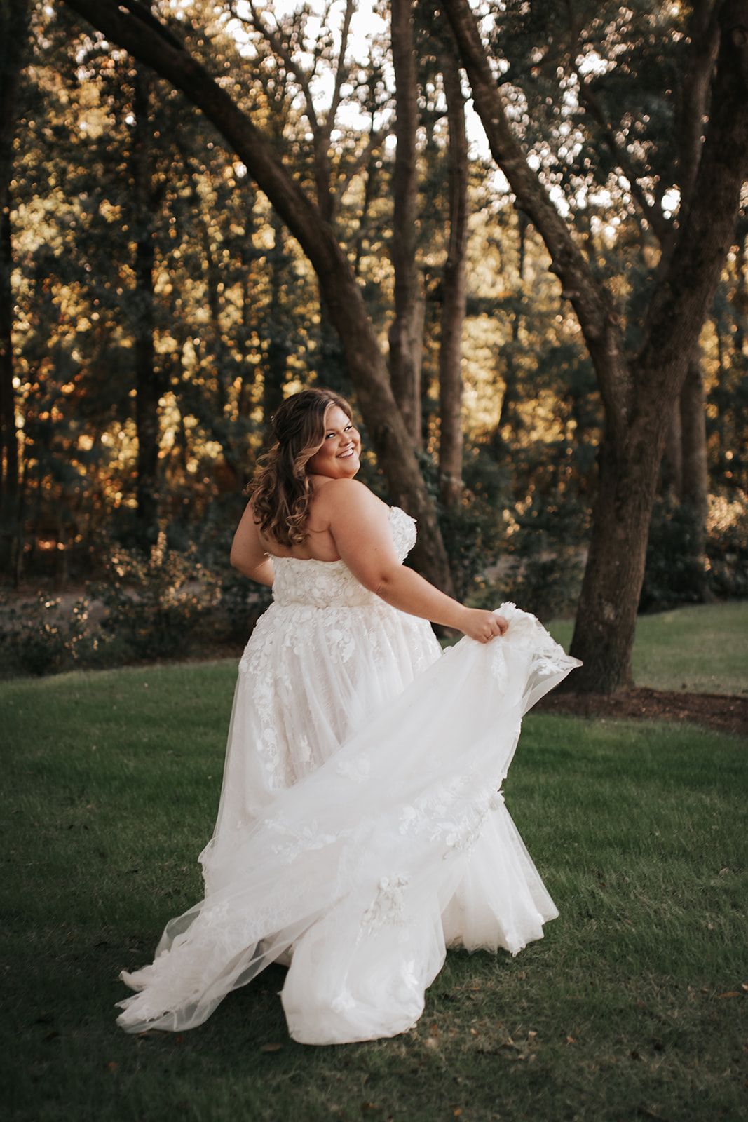 Woman in a white flowing dress twirling in a grassy wooded area at sunset