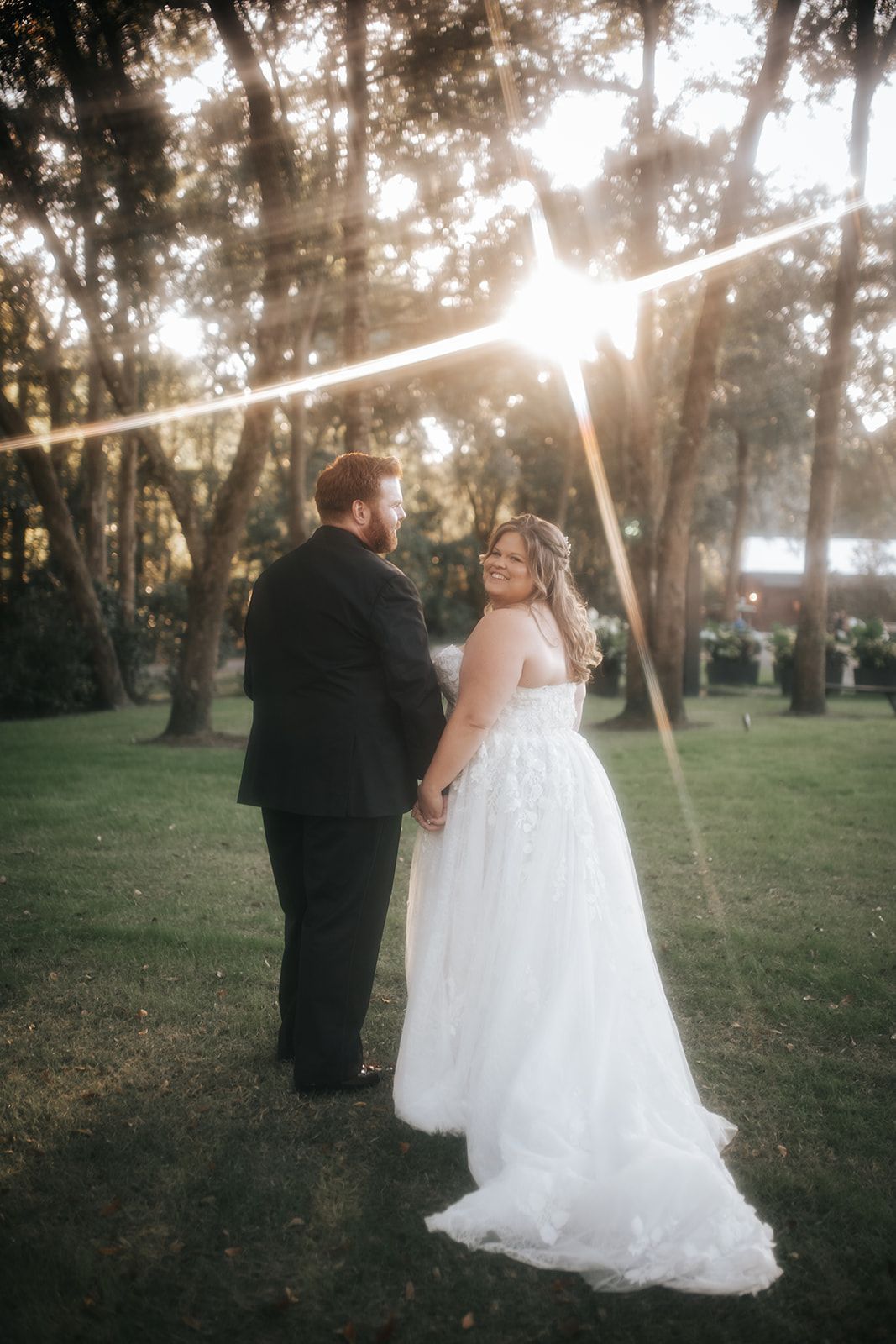 Newlyweds standing together in a sunlit park, bride in white gown and groom in dark suit, facing away under trees