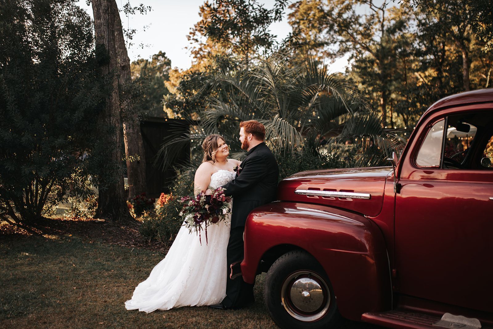 Bride and groom standing beside a vintage red truck in a wooded outdoor setting