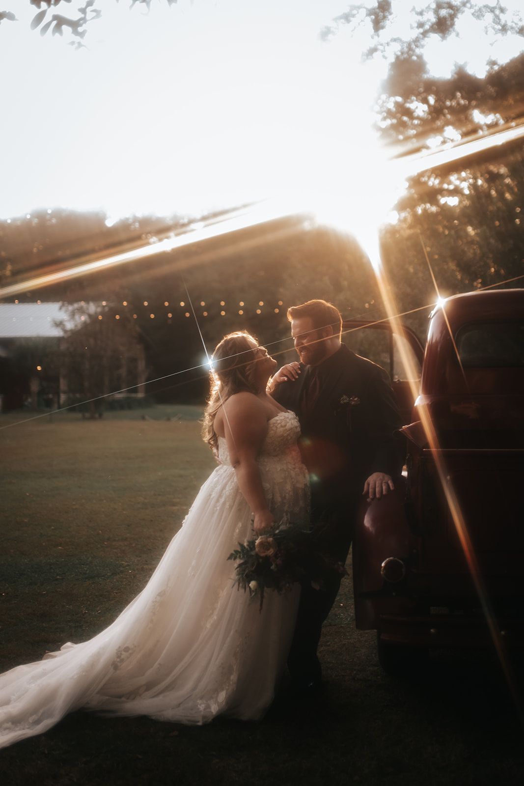 Bride and groom embrace at sunset outdoors under string lights, with a wedding gown train flowing behind them