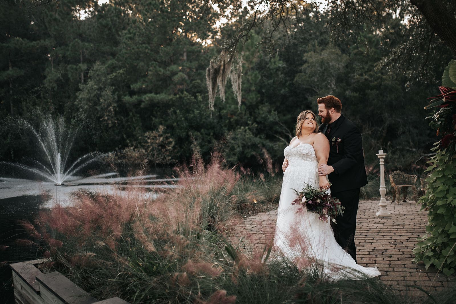 Couple in wedding attire standing by a fountain in a lush garden at dusk