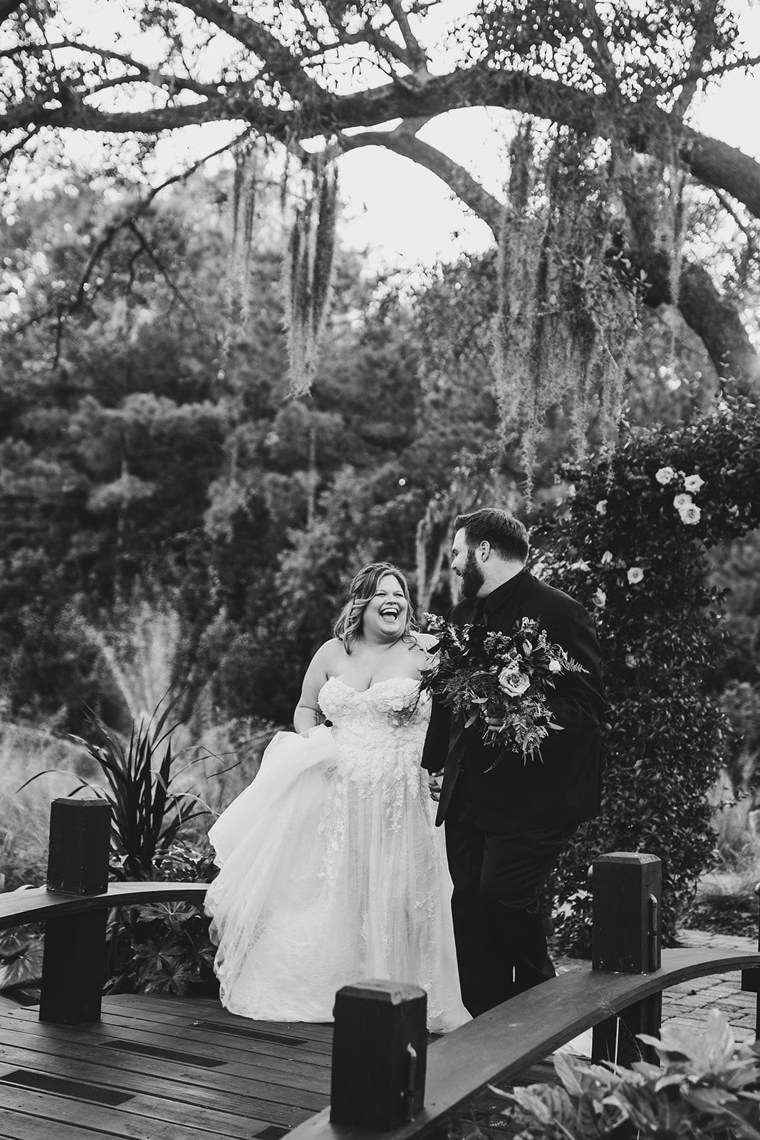 Bride and groom pose in a garden beneath arching trees, the bride holding a bouquet.
