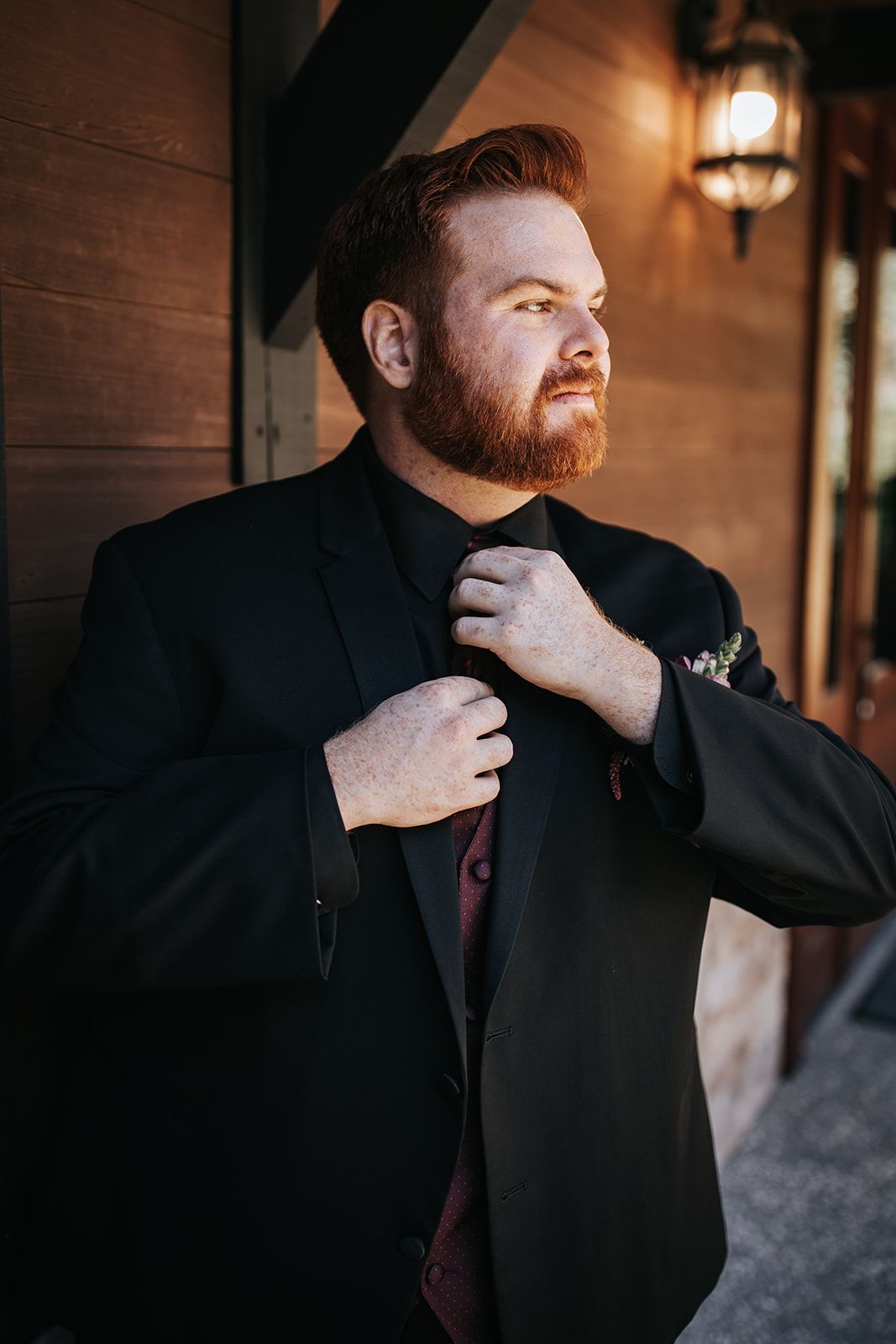 Man adjusting his tie in a black suit beside a wooden wall and warm lamp lighting
