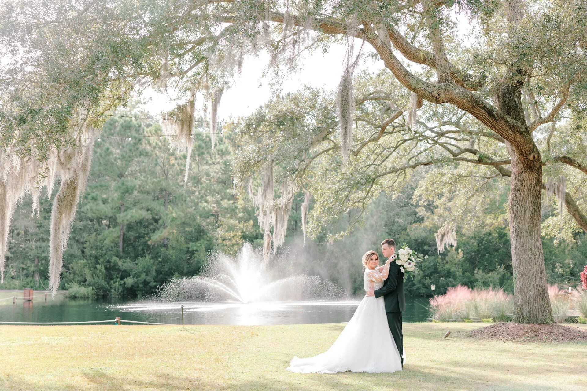 Bride and groom in a garden under mossy trees, dancing beside a fountain
