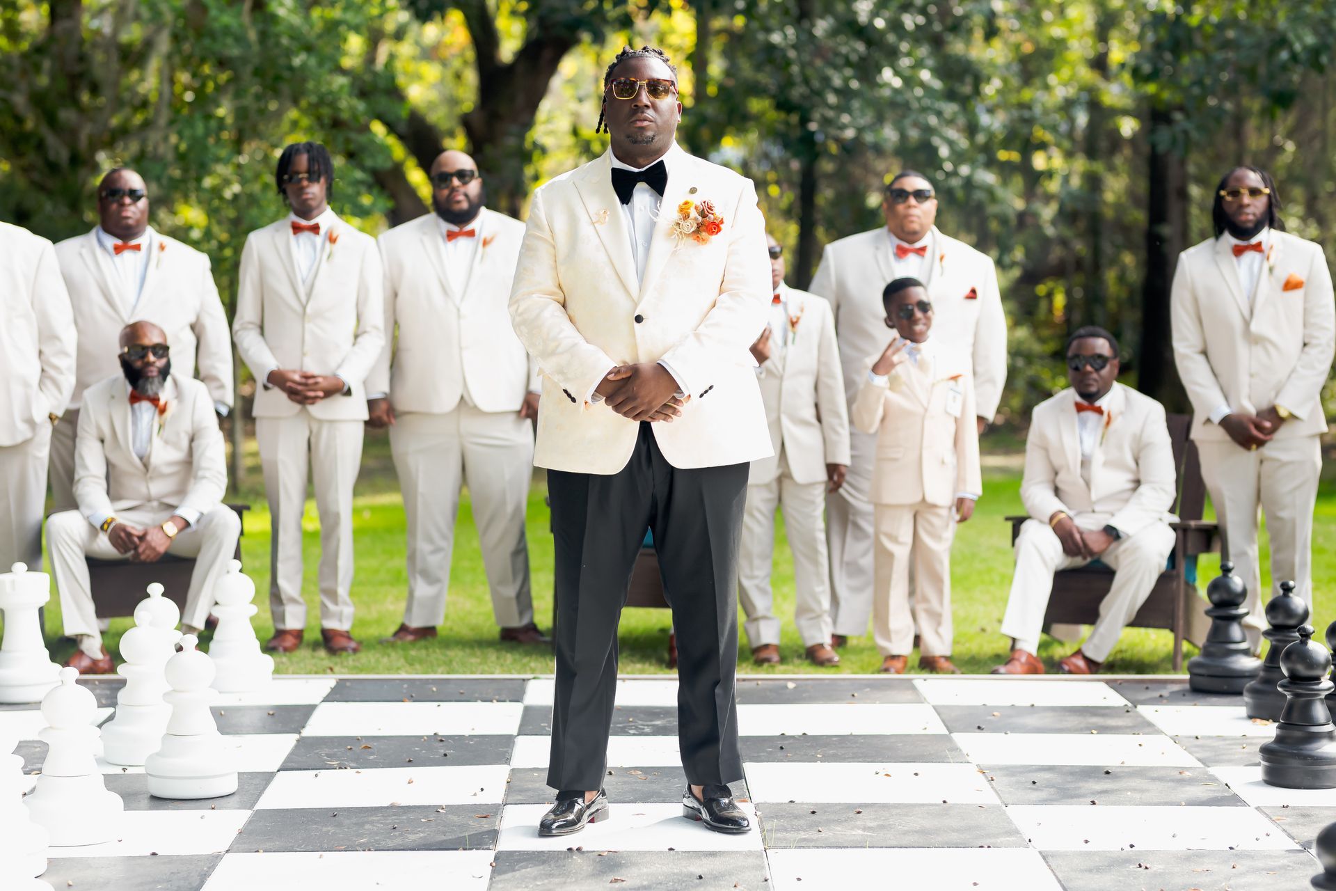 Groomsmen in cream suits pose on a giant chessboard outdoors, with the groom centered in front.