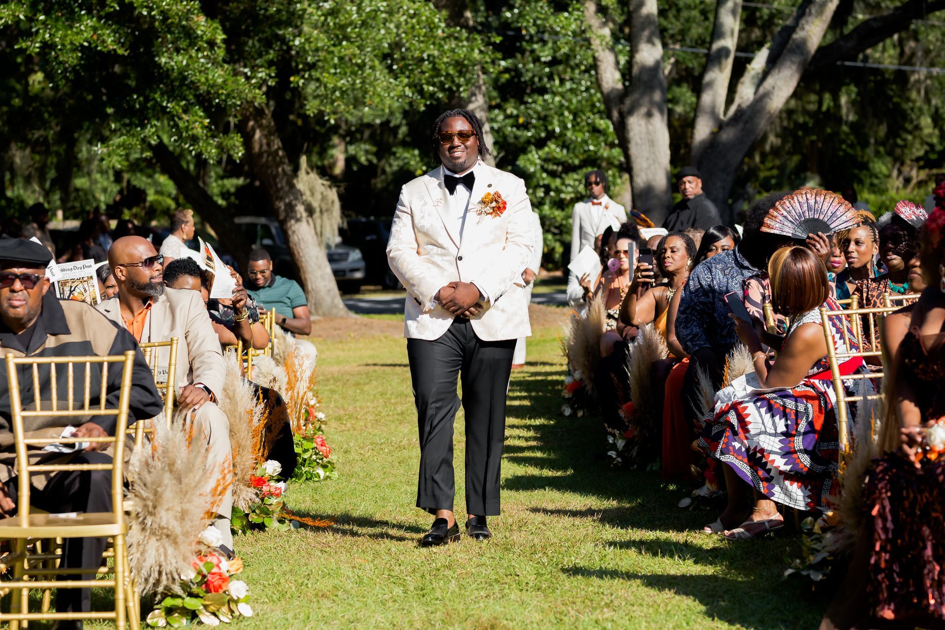 Man standing in a white suit holding a bouquet at an outdoor ceremony with seated guests