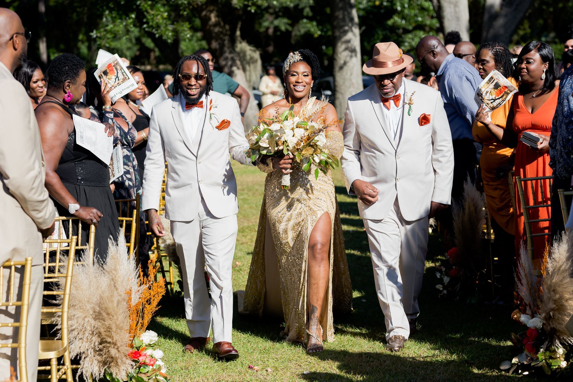 Wedding party walking outdoors, with a bride in a gold dress and groom in a white suit among guests.