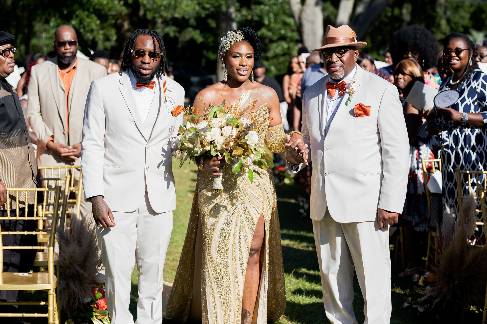 Wedding party walking outdoors, with bride in gold gown and groom in white suit, holding hands in a garden.
